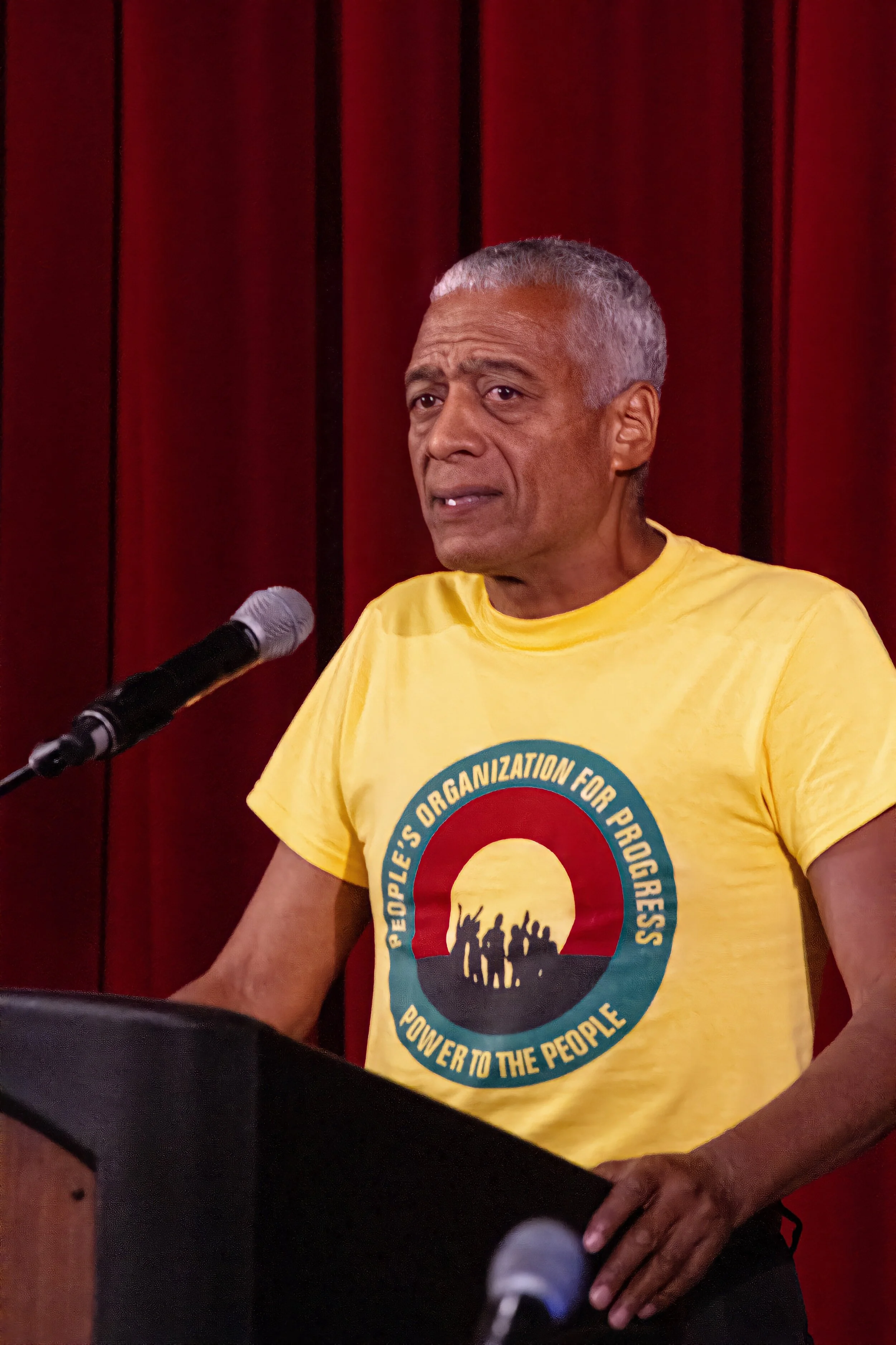 An older man with gray hair speaking at a podium with a microphone, wearing a yellow T-shirt with a circular logo featuring silhouettes of people and text.