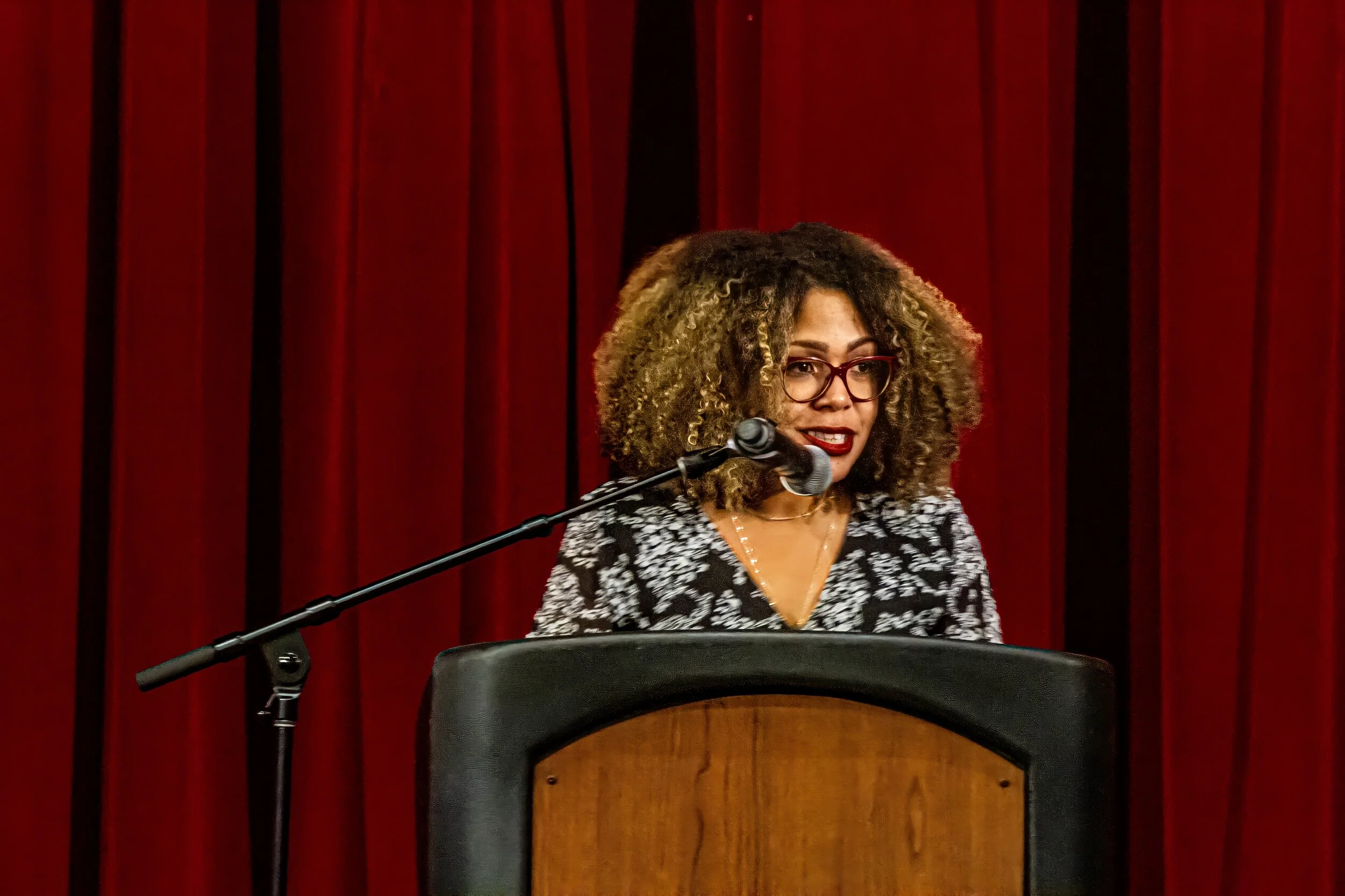 A woman with curly hair, glasses, and a patterned top stands behind a podium with a microphone, in front of red curtains.