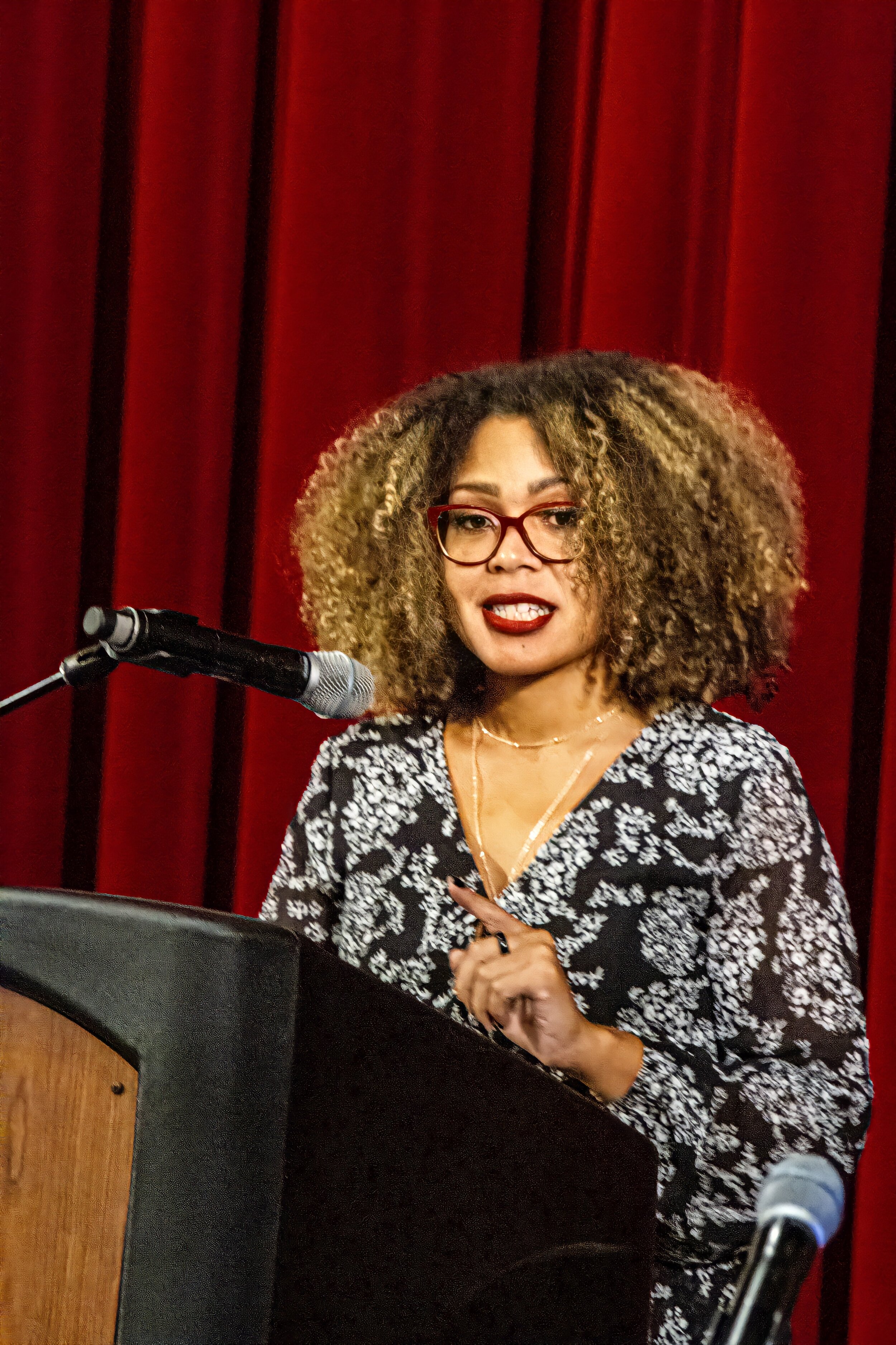 Woman with curly blonde hair and red glasses speaking at a podium in front of red curtains.