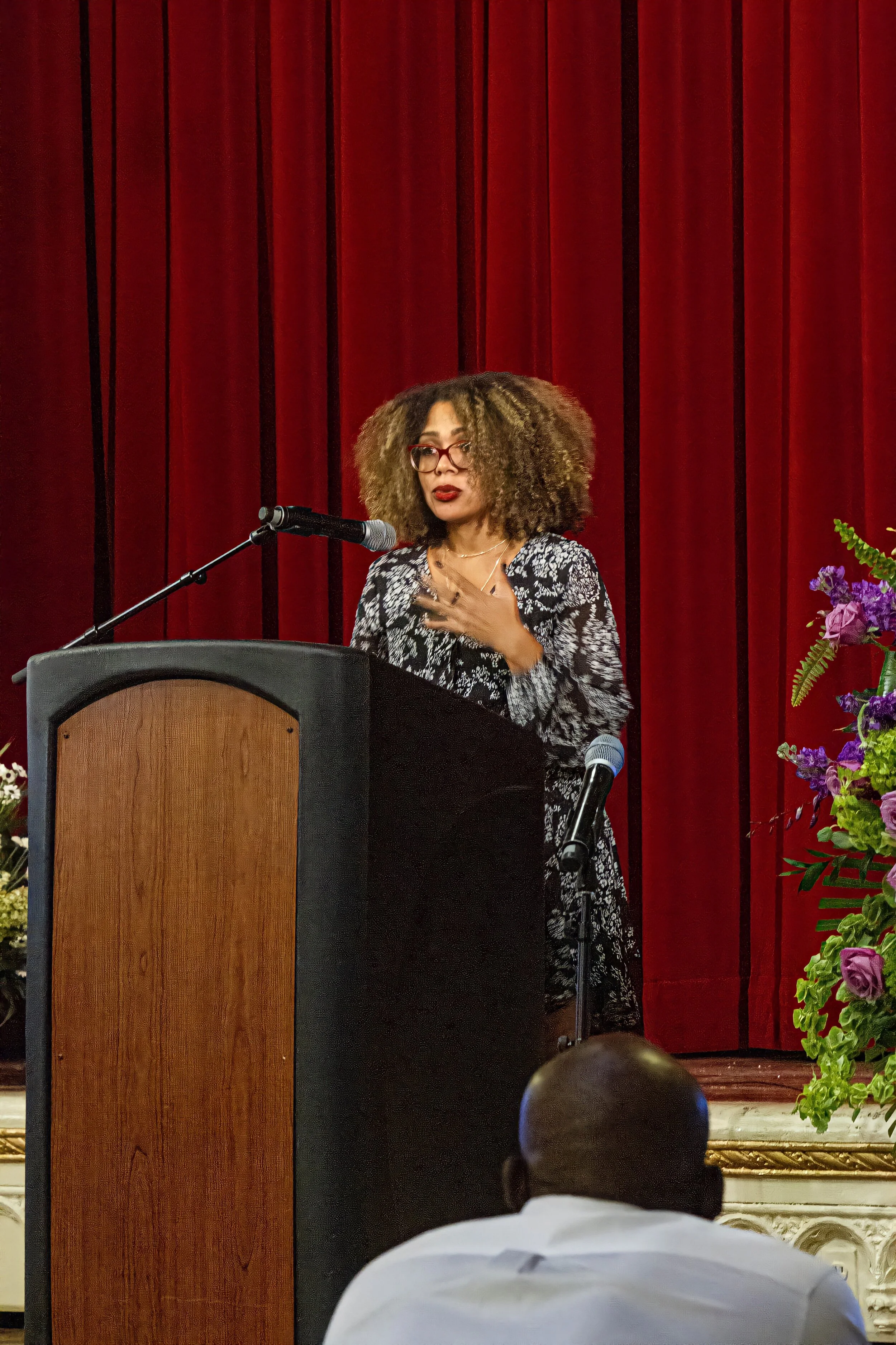 A woman with curly hair and glasses is speaking at a podium with a microphone, in front of a red curtain, while an audience member in a white shirt is visible in the foreground.