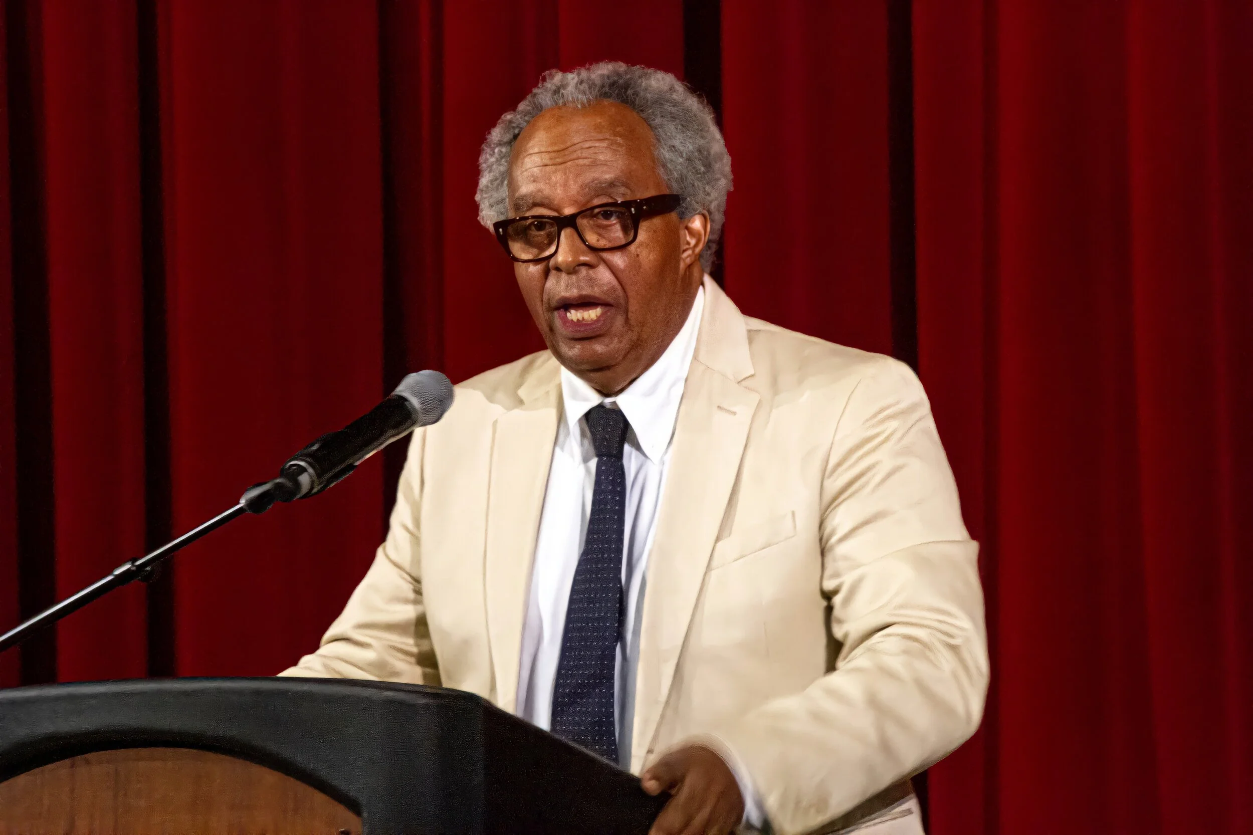 An elderly man with gray hair, glasses, and a beige suit speaking at a podium in front of red curtains.