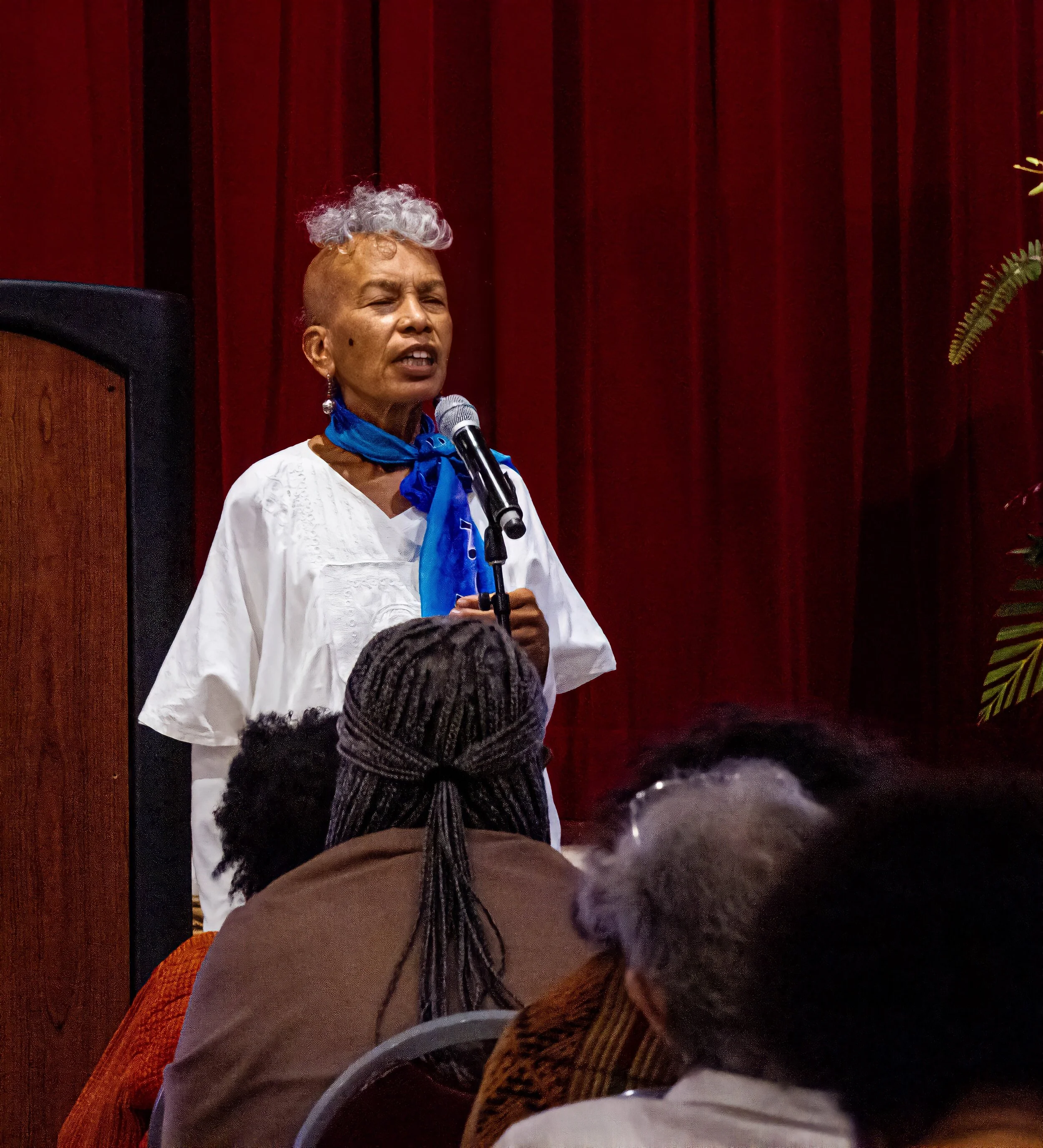 A woman with short hair and grey curls on top, wearing a white blouse and a blue scarf, is speaking into a microphone in front of a red curtain, with plants visible on the side.