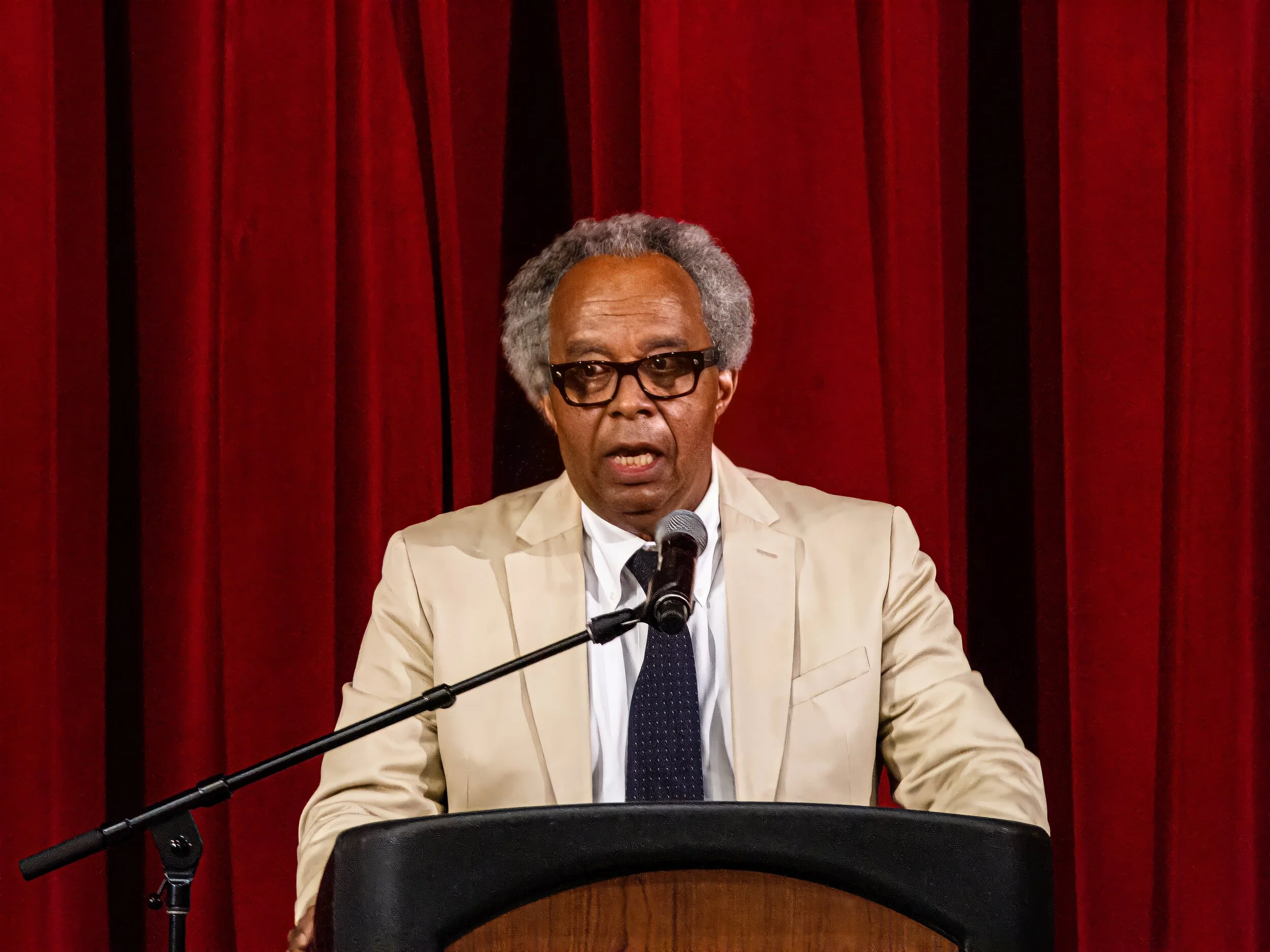 An older man with gray hair and glasses, wearing a beige suit jacket, white shirt, and dark tie, speaking at a podium with a microphone in front of a red curtain.