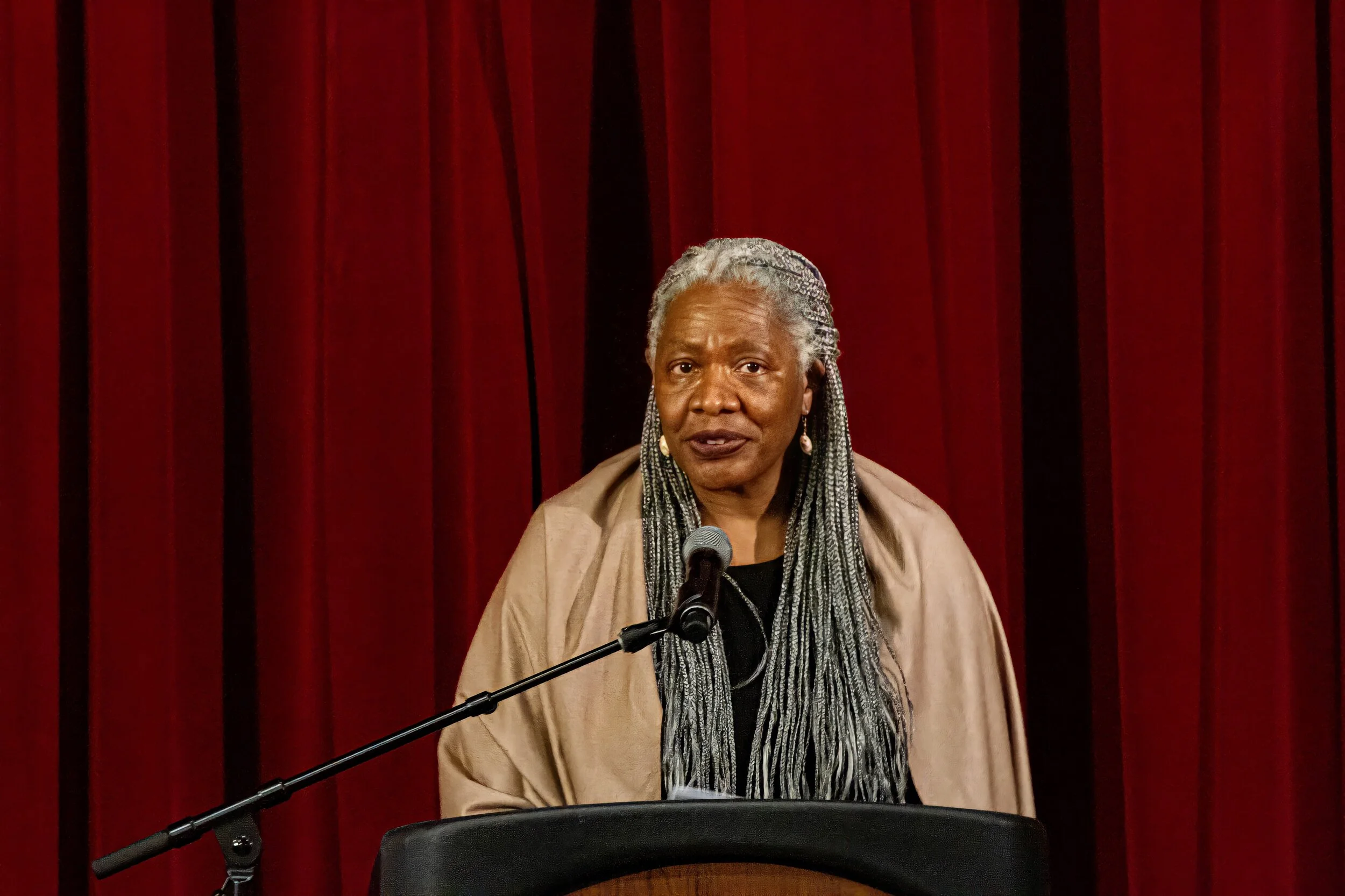 An older African American woman with long gray braided hair speaking at a podium with a microphone against a red curtain background.