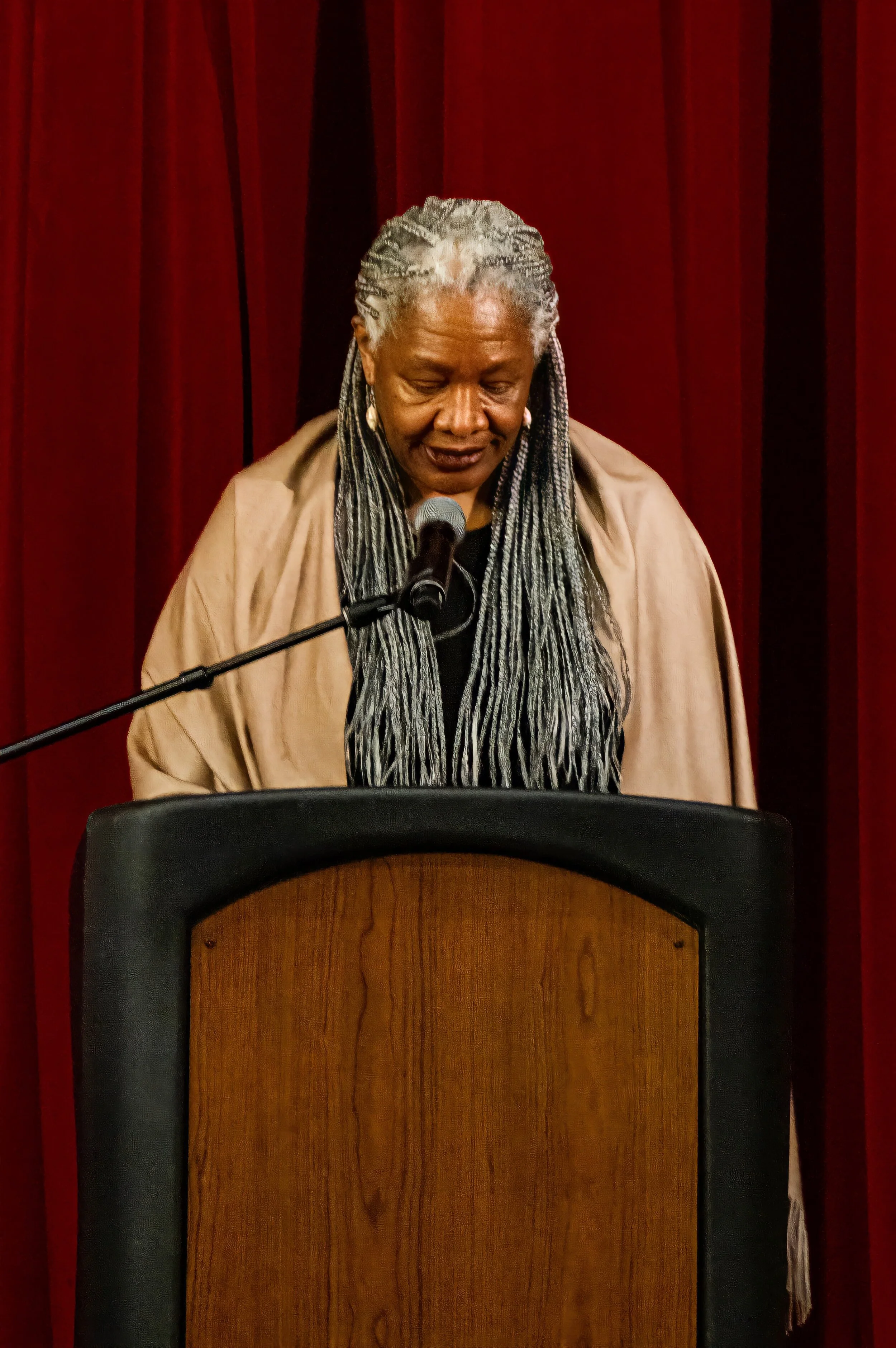 An African American woman with gray braided hair wearing a beige shawl, speaking or reading into a microphone at a lectern with red curtains behind her.