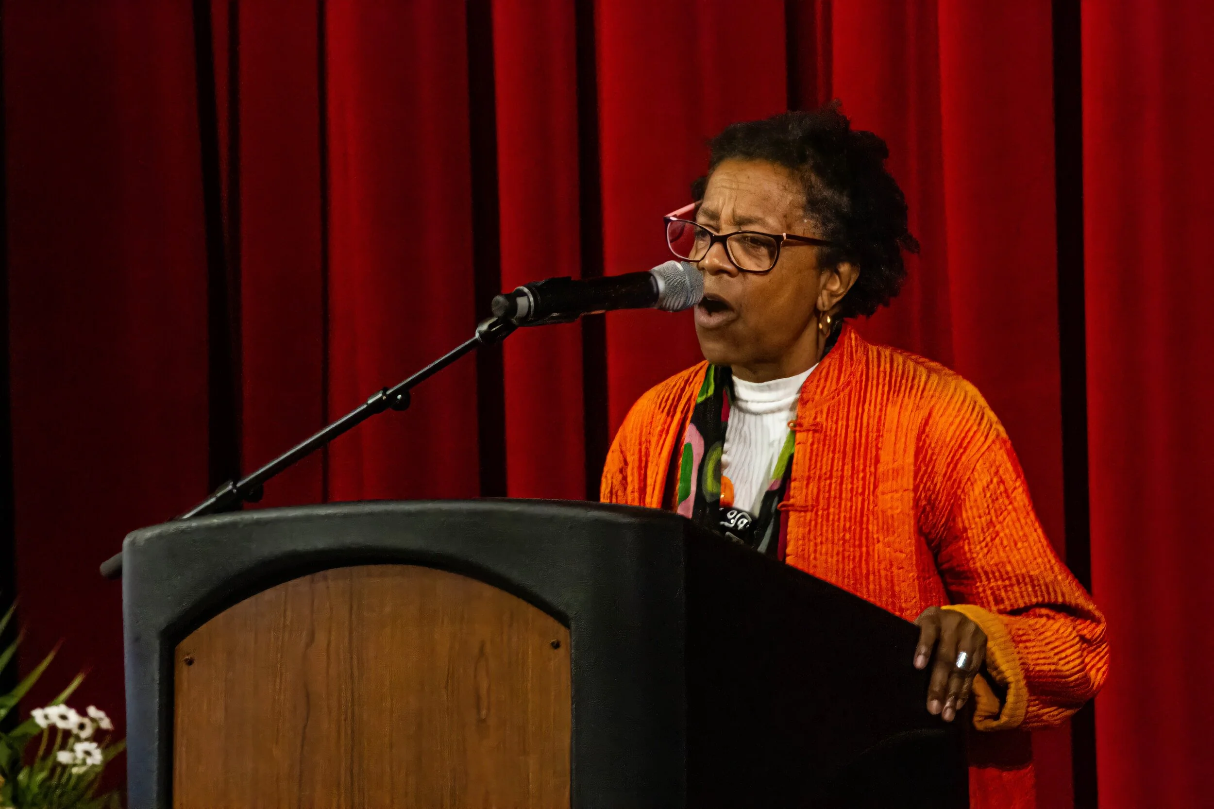 Woman speaking at a podium with a microphone, wearing glasses and an orange jacket, against a red curtain background.