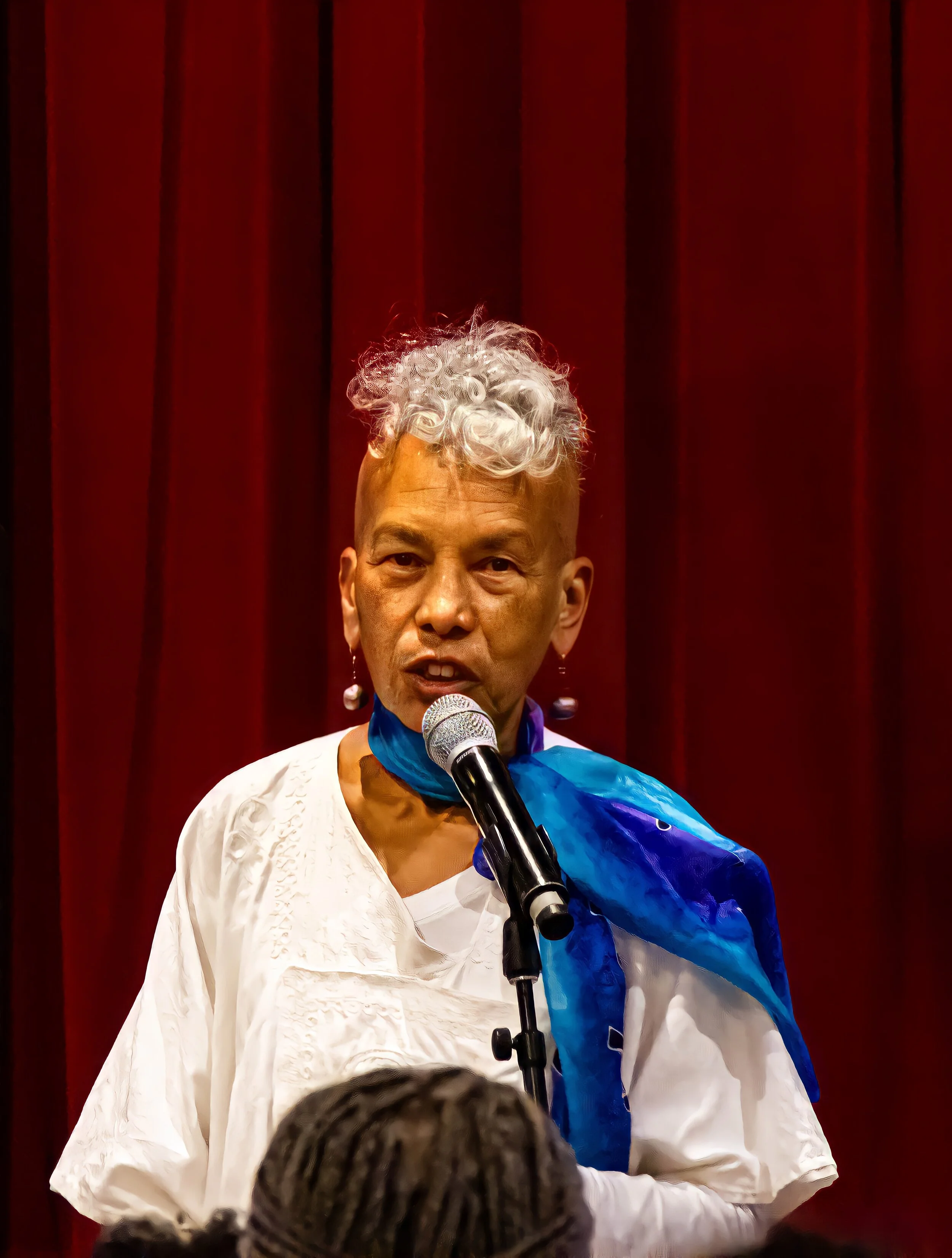 A woman with silver curly hair speaking into a microphone, wearing a white embroidered dress and a blue cape, standing in front of red curtains.