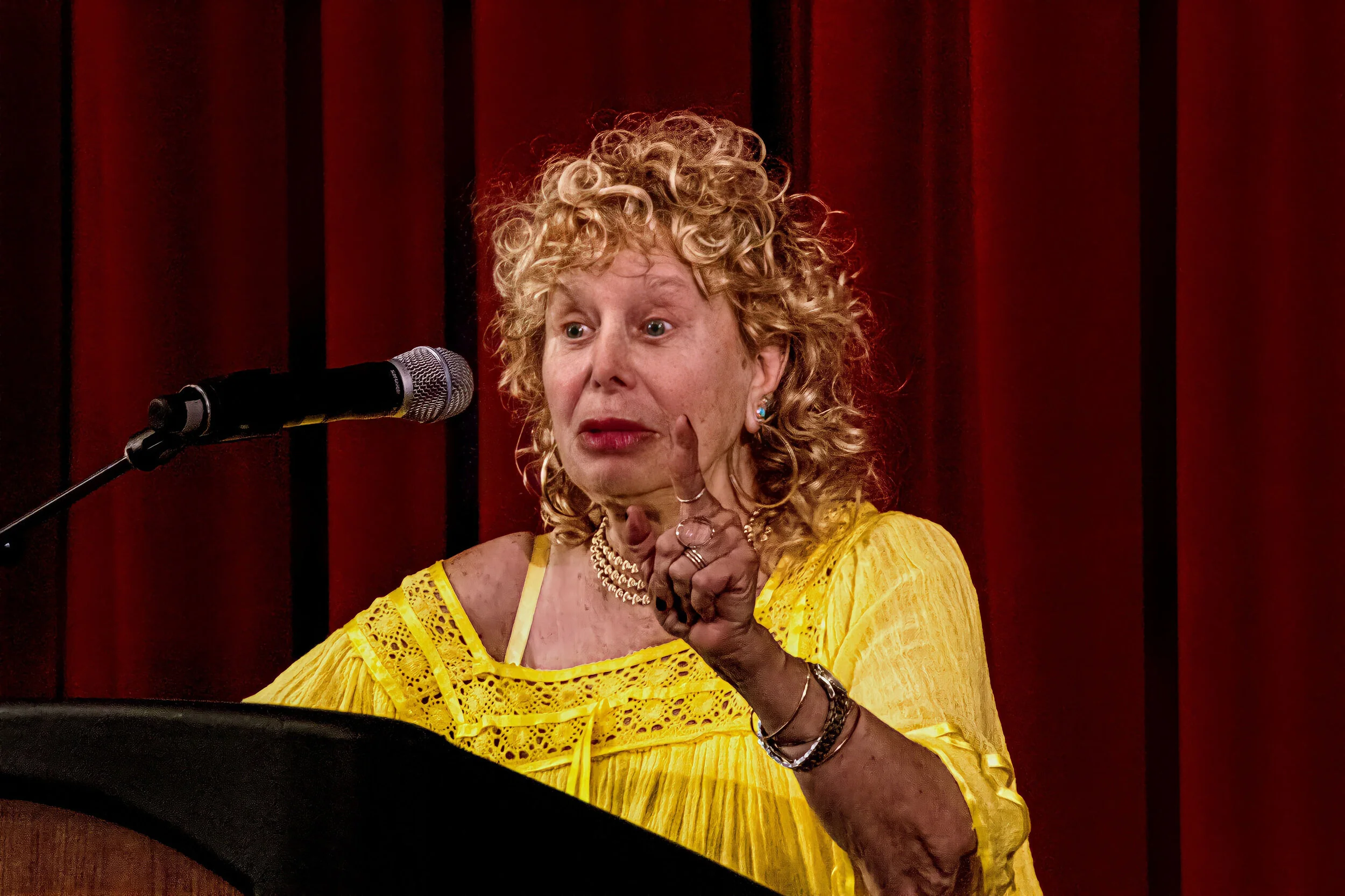 An older woman with curly blonde hair wearing a yellow lace dress is speaking into a microphone on a stage with red curtains. She has jewelry including rings, bracelets, earrings, and a necklace, and is gesturing with her hand.