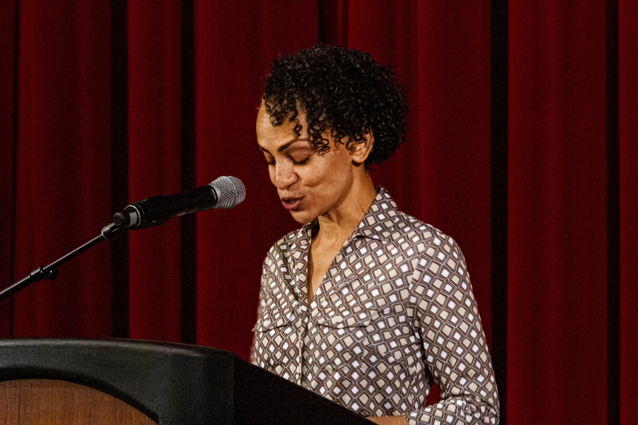 A woman with short curly hair wearing a patterned shirt singing into a microphone.