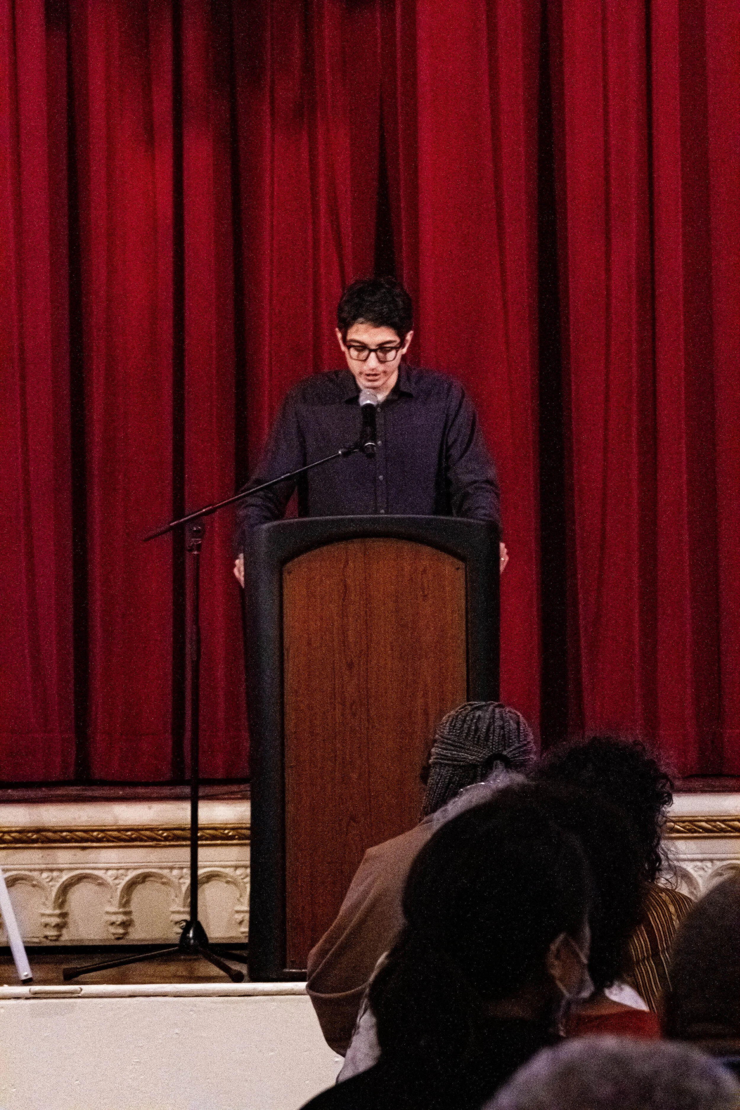 A young man with glasses standing at a podium on stage, speaking into a microphone, with red curtains in the background, during a presentation or speech.
