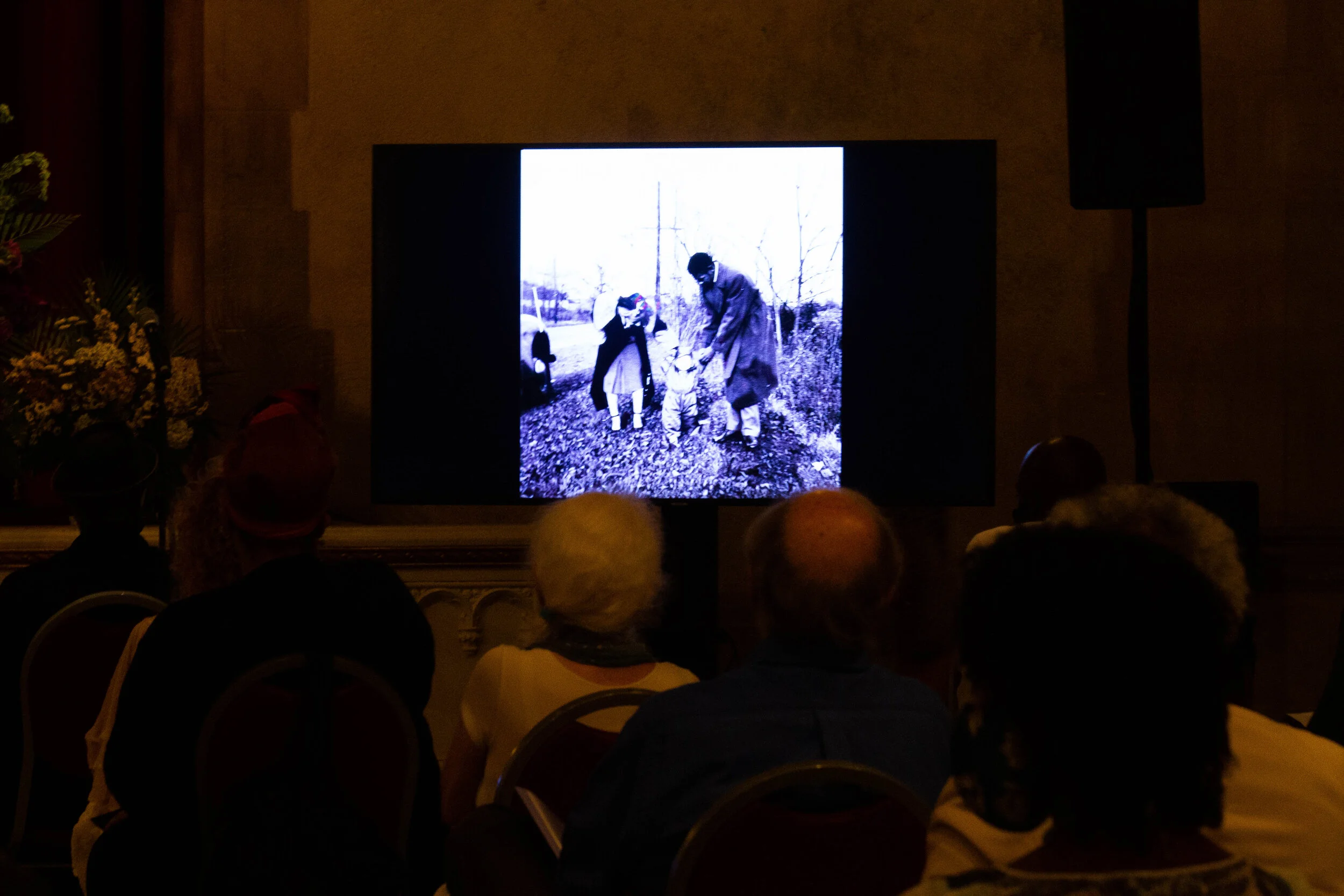 A black-and-white photograph projected on a screen showing children planting trees, with adults supervising in a rural outdoor setting.