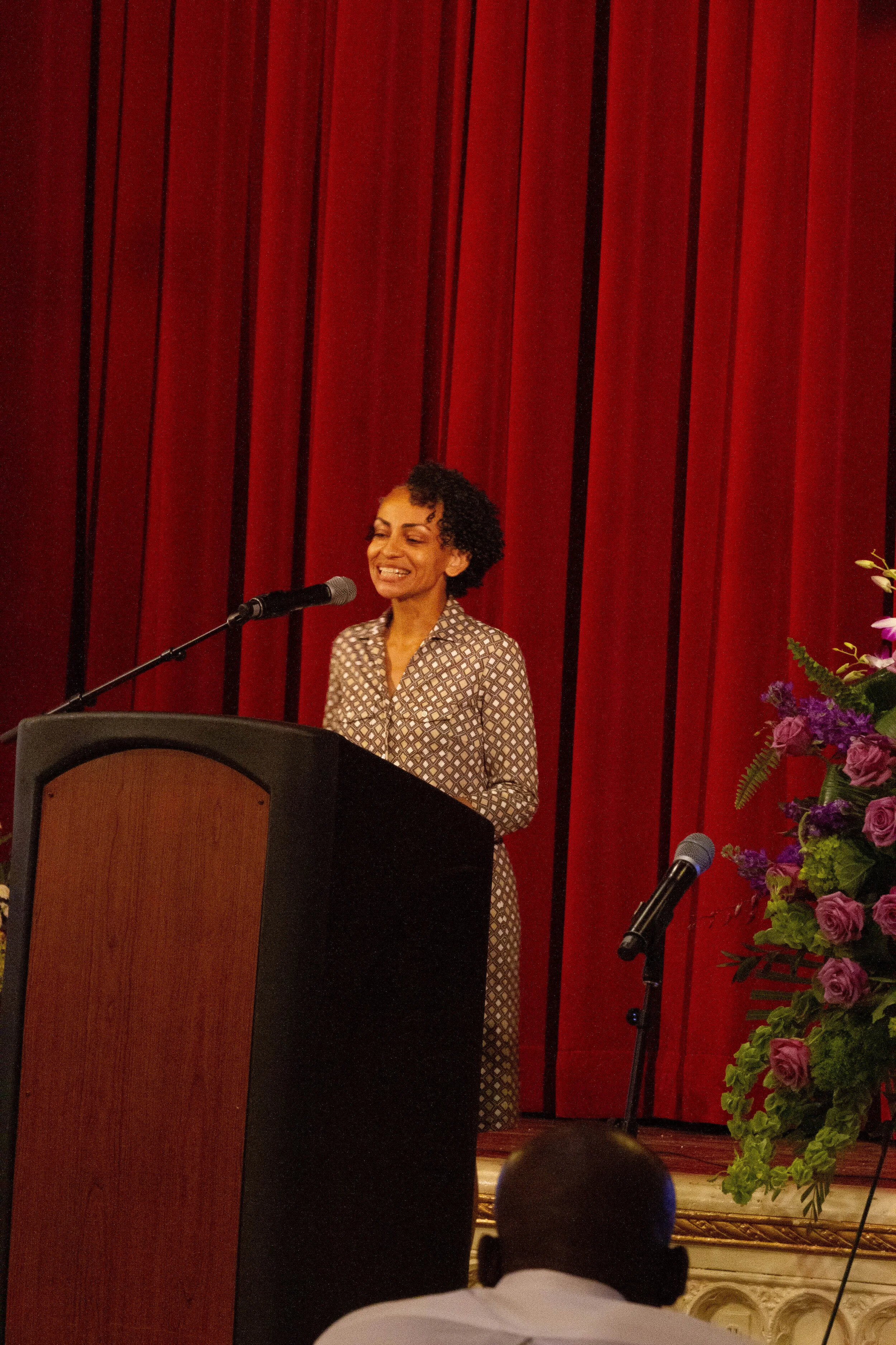 A woman with curly hair smiling while speaking at a podium in front of a red curtain, with a large bouquet of purple and green flowers to her right.