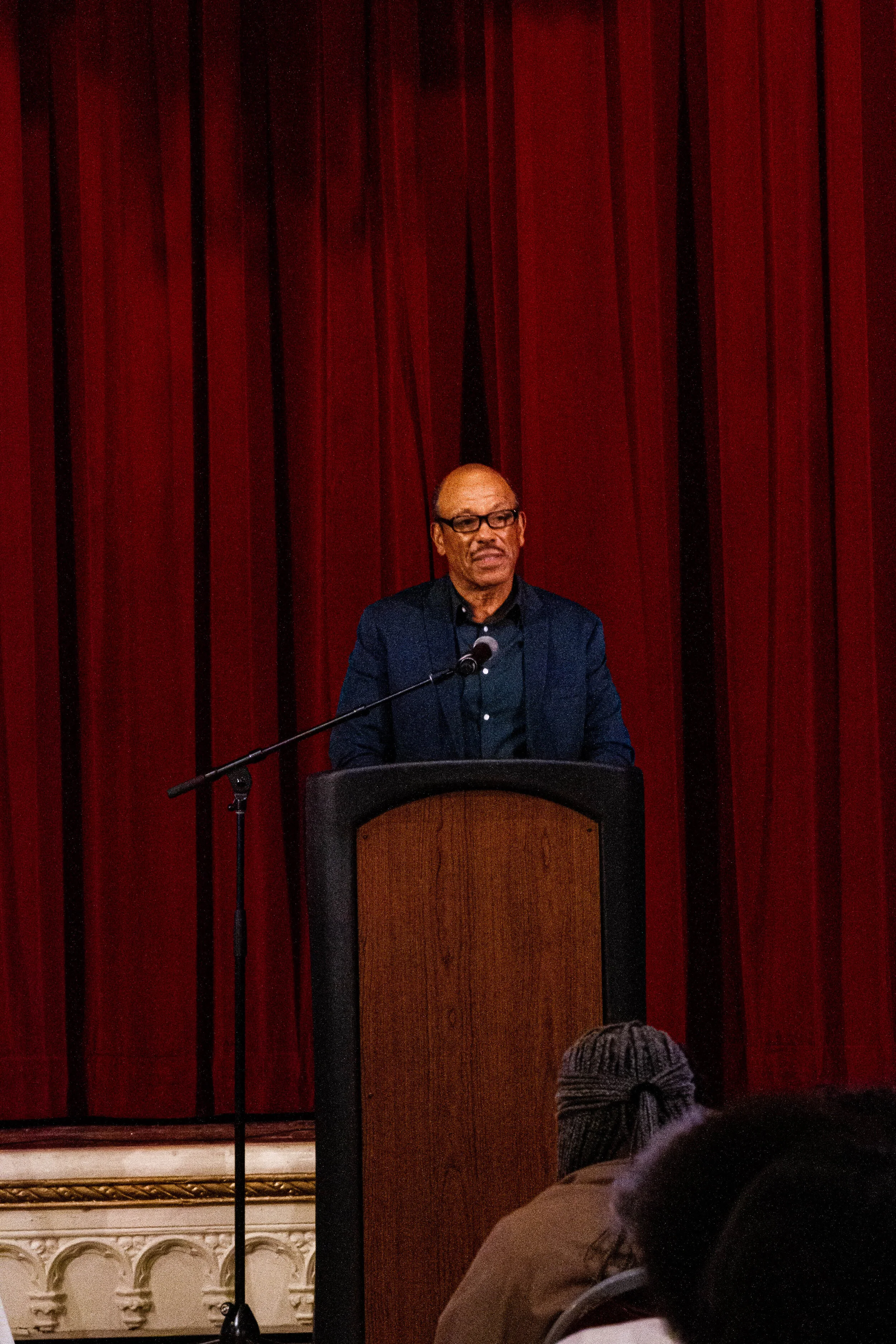 A man with glasses and a dark blazer is standing at a podium in front of red curtains, speaking into a microphone. An audience is visible in the foreground.