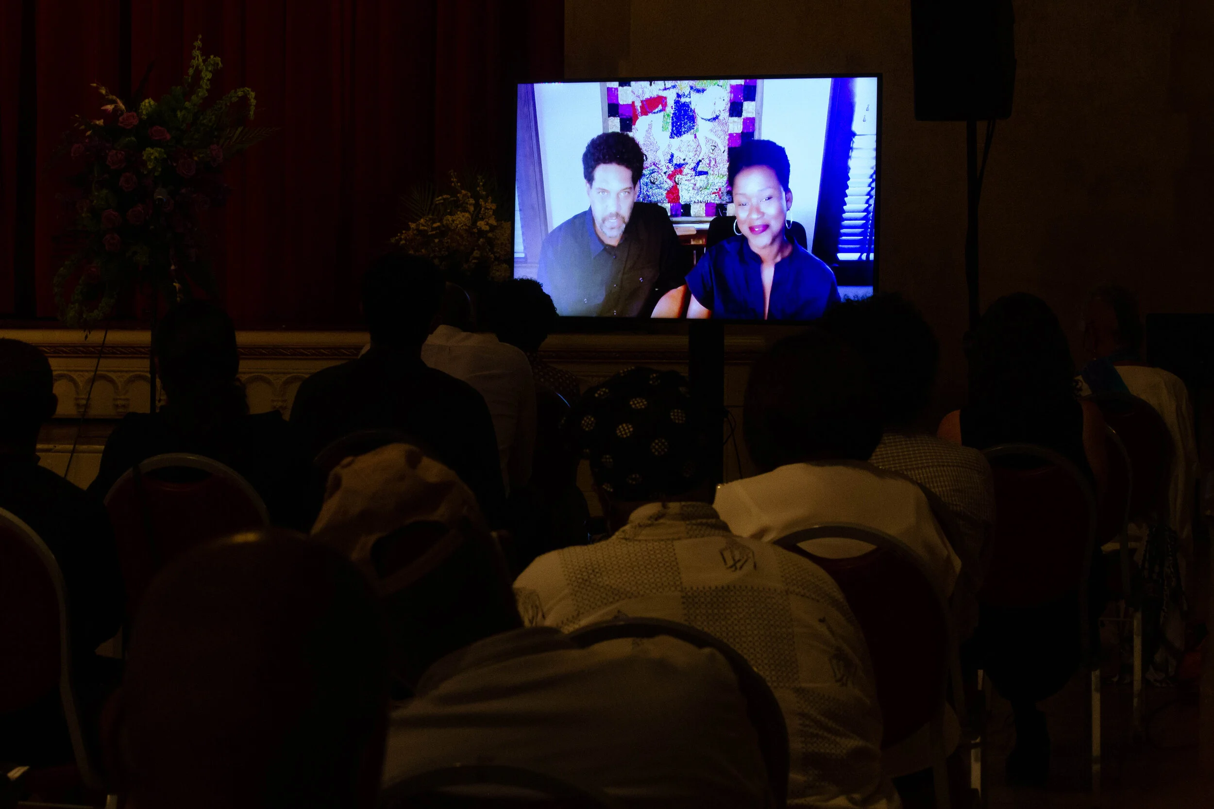 Audience viewing a large screen showing two people in a video call at an indoor event, with a floral arrangement on the side.