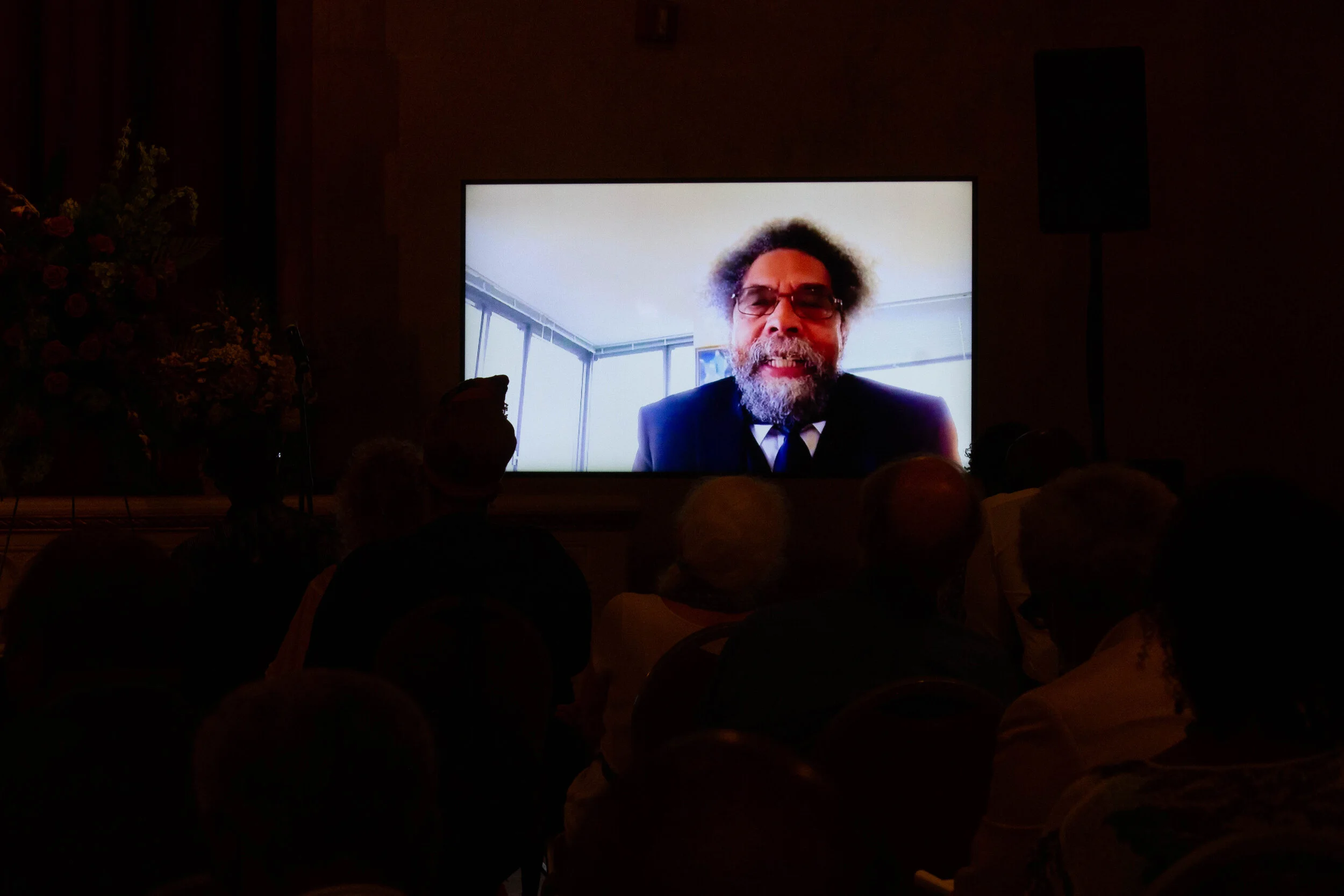 Man speaking on a video call displayed on a large screen, audience in silhouette in a dimly lit conference room.