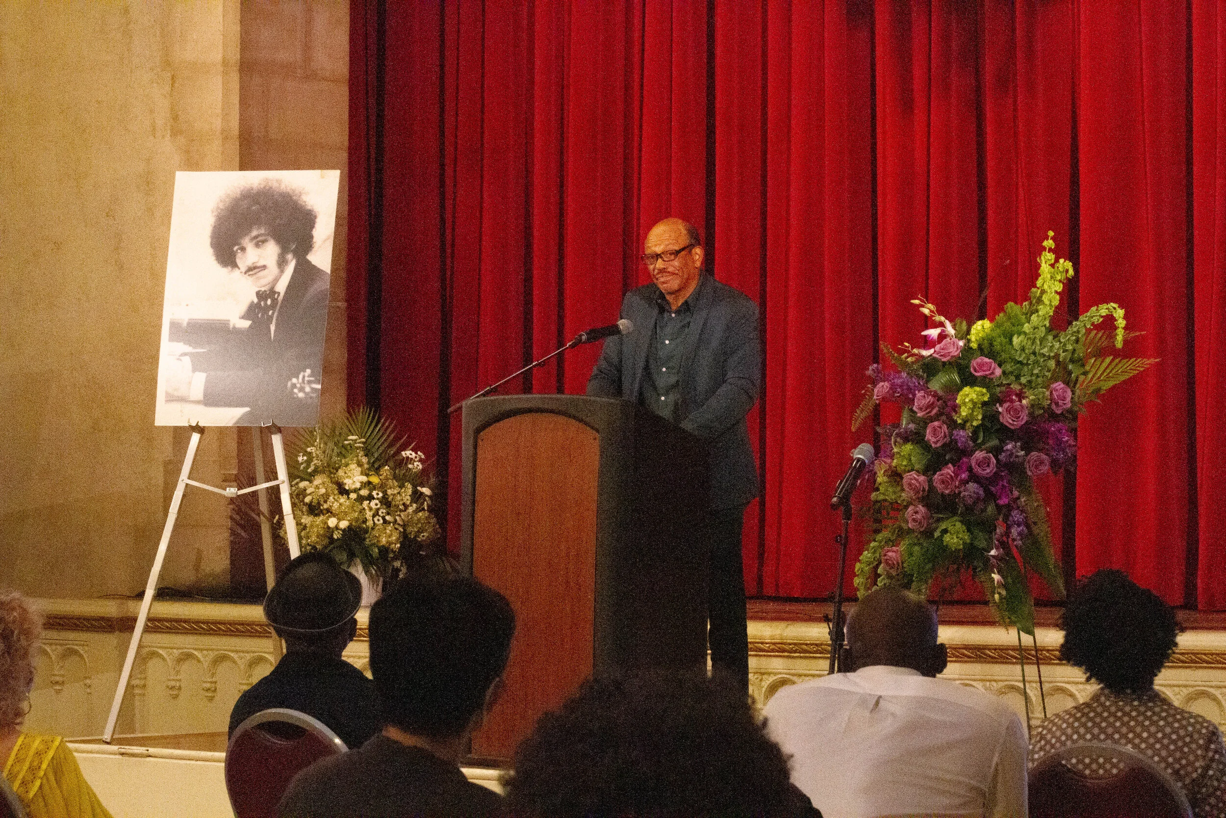 A man speaking at a podium during a memorial event, with a large portrait of a man with an afro hairstyle on an easel to his left, and a tall bouquet of purple, green, and yellow flowers to his right, red curtain backdrop behind him, and audience mem