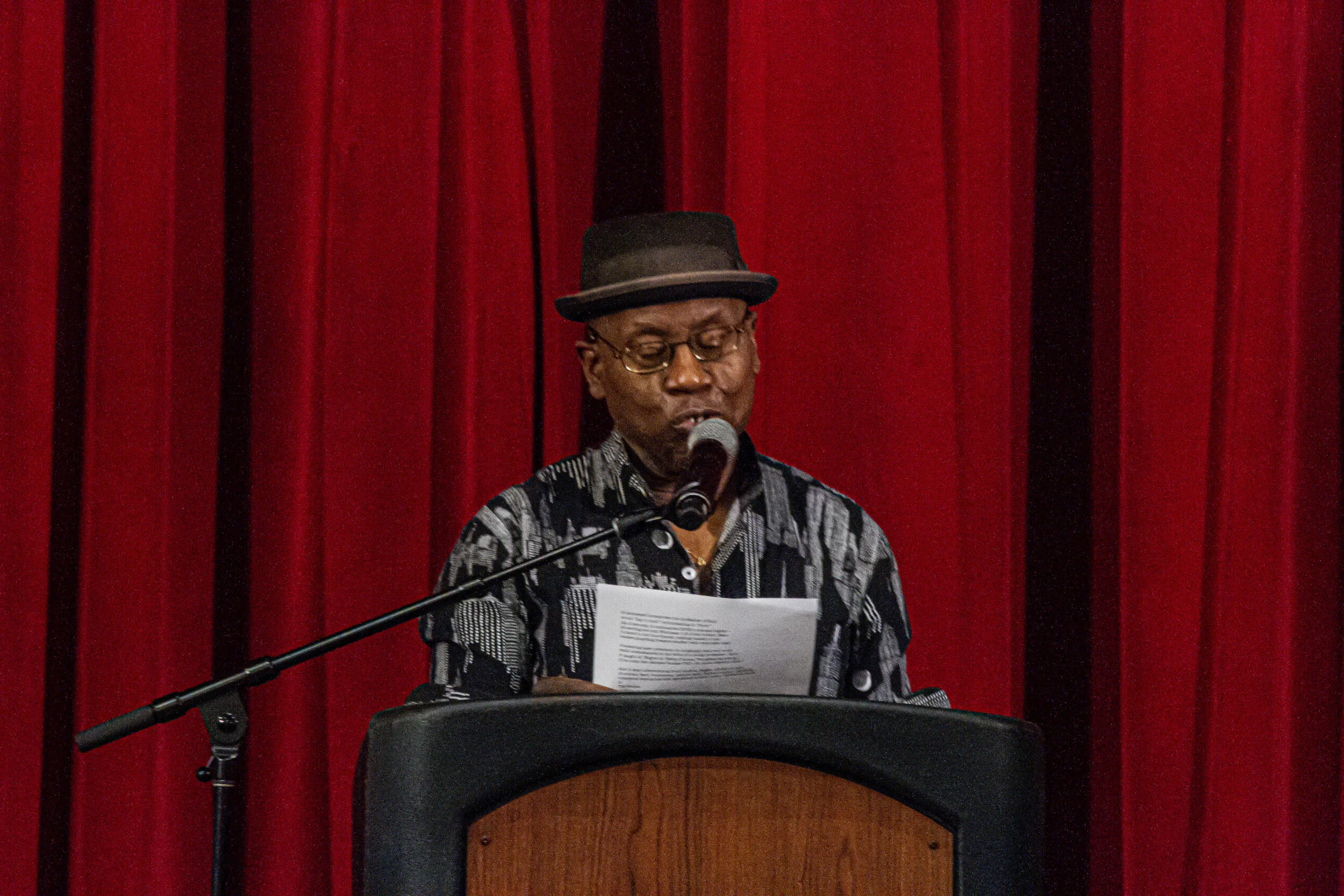 An elderly woman wearing glasses and a black hat reads from a paper while standing at a podium with a microphone, in front of a red curtain background.