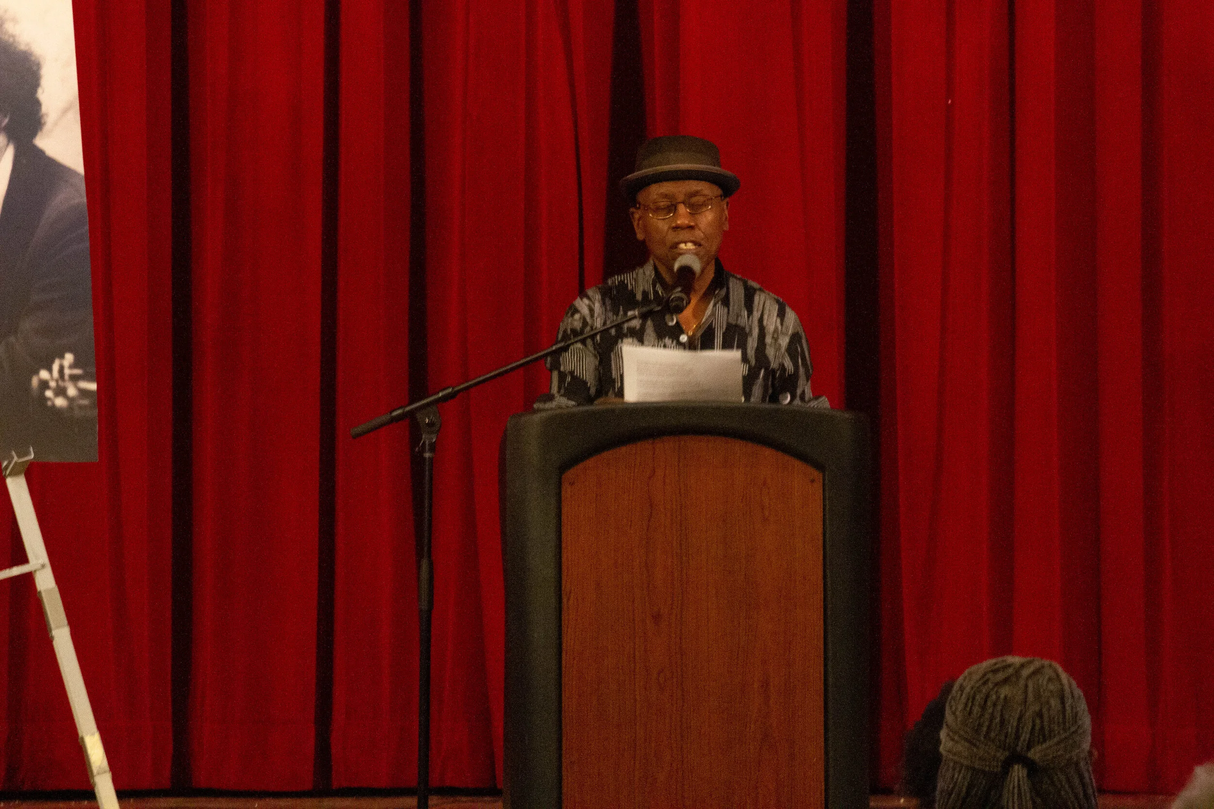 A person wearing glasses and a hat stands at a podium with a microphone and reads from a paper in front of red stage curtains.