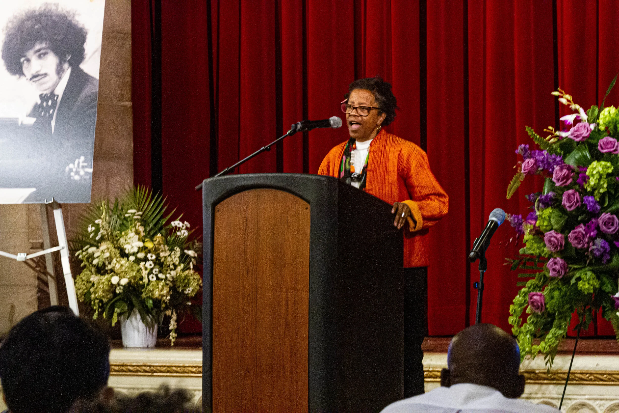 A woman with glasses and short curly hair, wearing an orange jacket, speaks at a podium during an event. There are large floral arrangements on either side of the stage and a portrait of a man with an afro hairstyle displayed on an easel to the left 