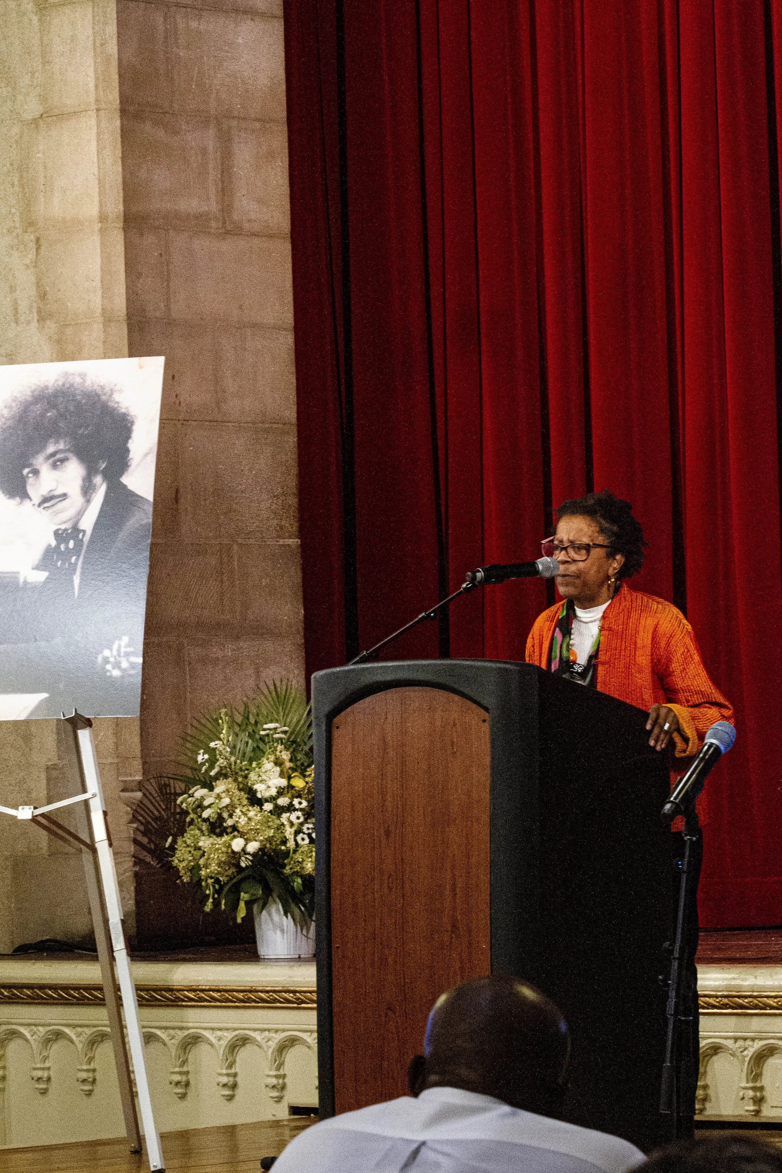 A woman speaking at a podium during a formal event, with a film poster and floral arrangement next to her, and an audience member in the foreground.