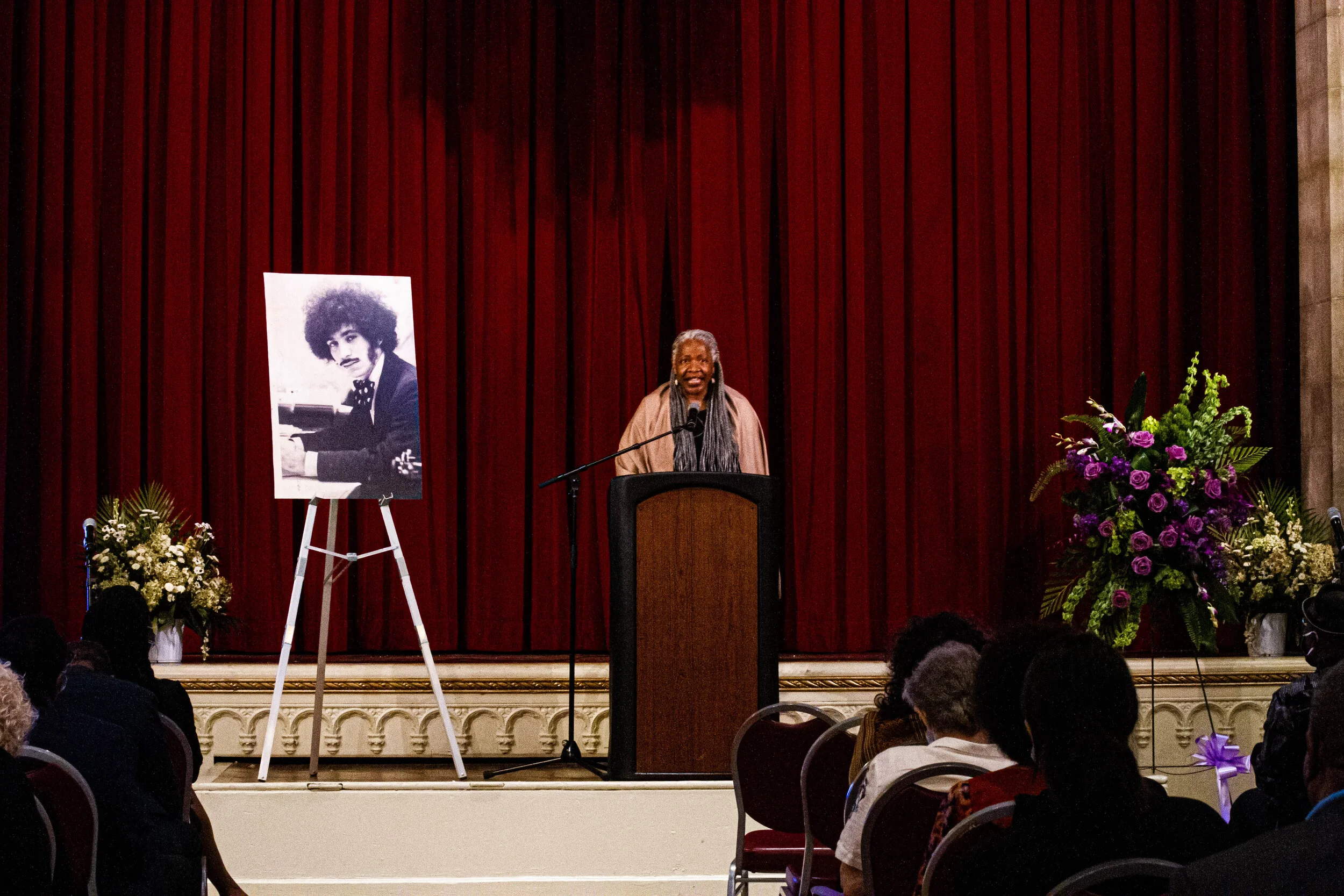 A woman is speaking at a podium during an event in a hall with a large red curtain in the background. To her left is a black and white portrait of a woman at a piano, on an easel. There are floral arrangements on both sides of the stage, including pu