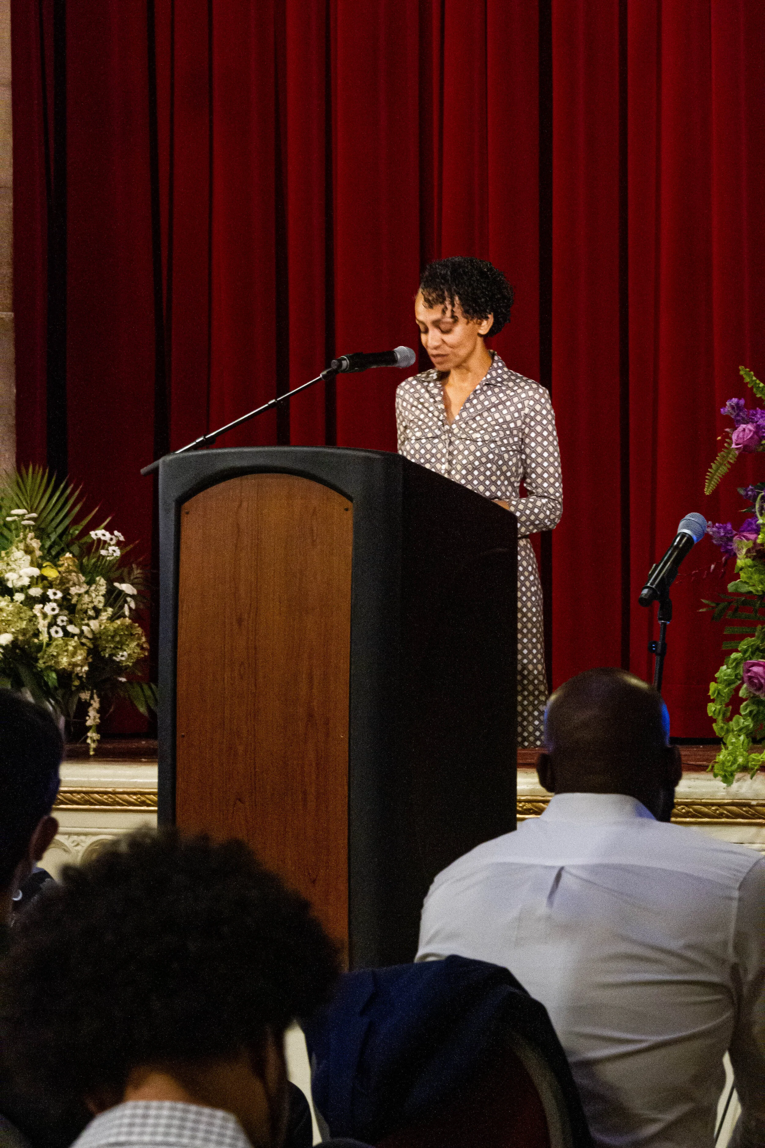 A woman with short curly hair is standing at a podium, speaking or reading, in front of a red curtain. There are flower arrangements on either side of the podium, and a microphone is positioned in front of her. Audience members are seated and listeni