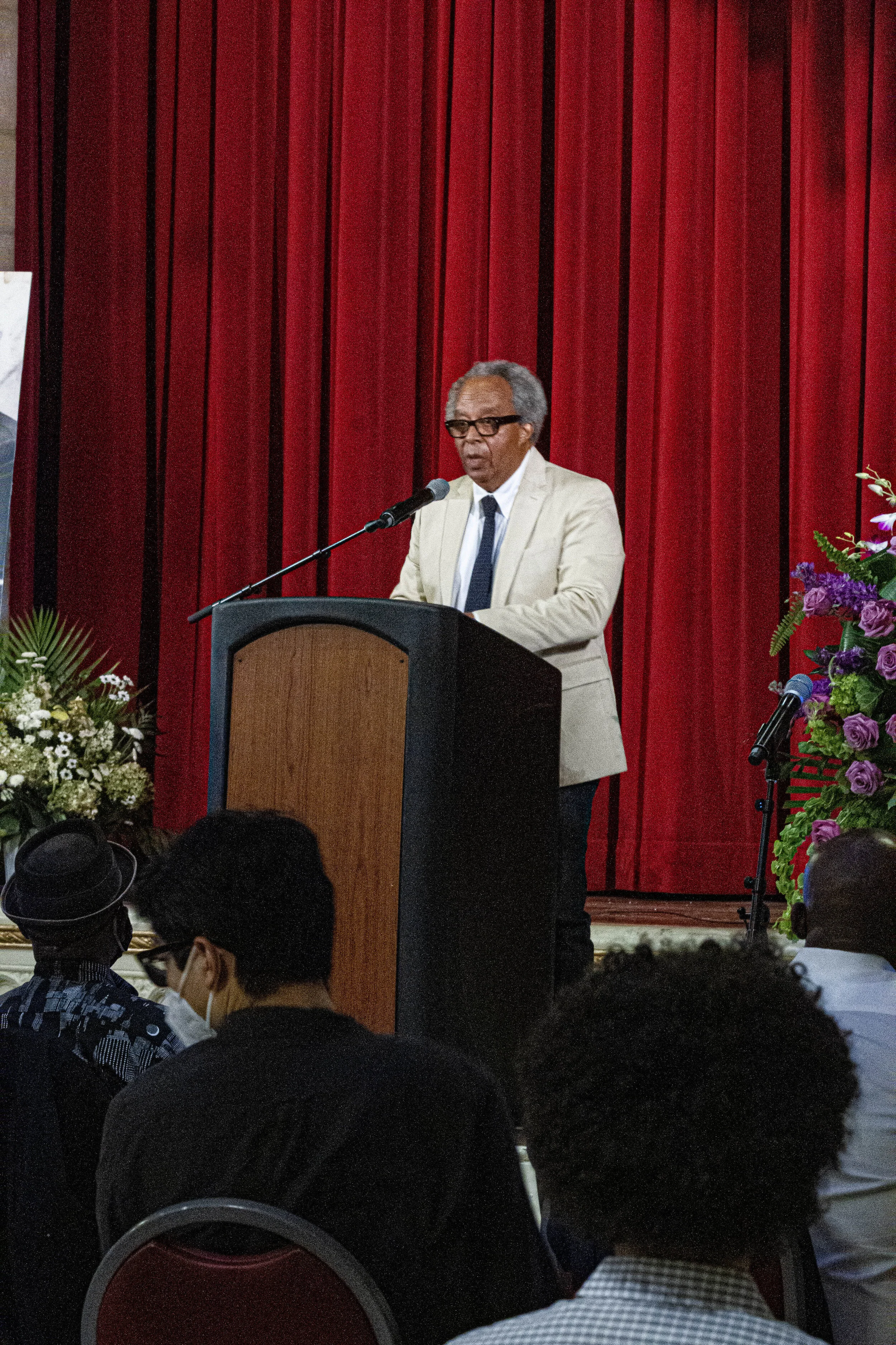 An elderly man in a beige suit and glasses is giving a speech at a podium with a microphone in front of a red curtain, with floral arrangements on either side, during a formal event.
