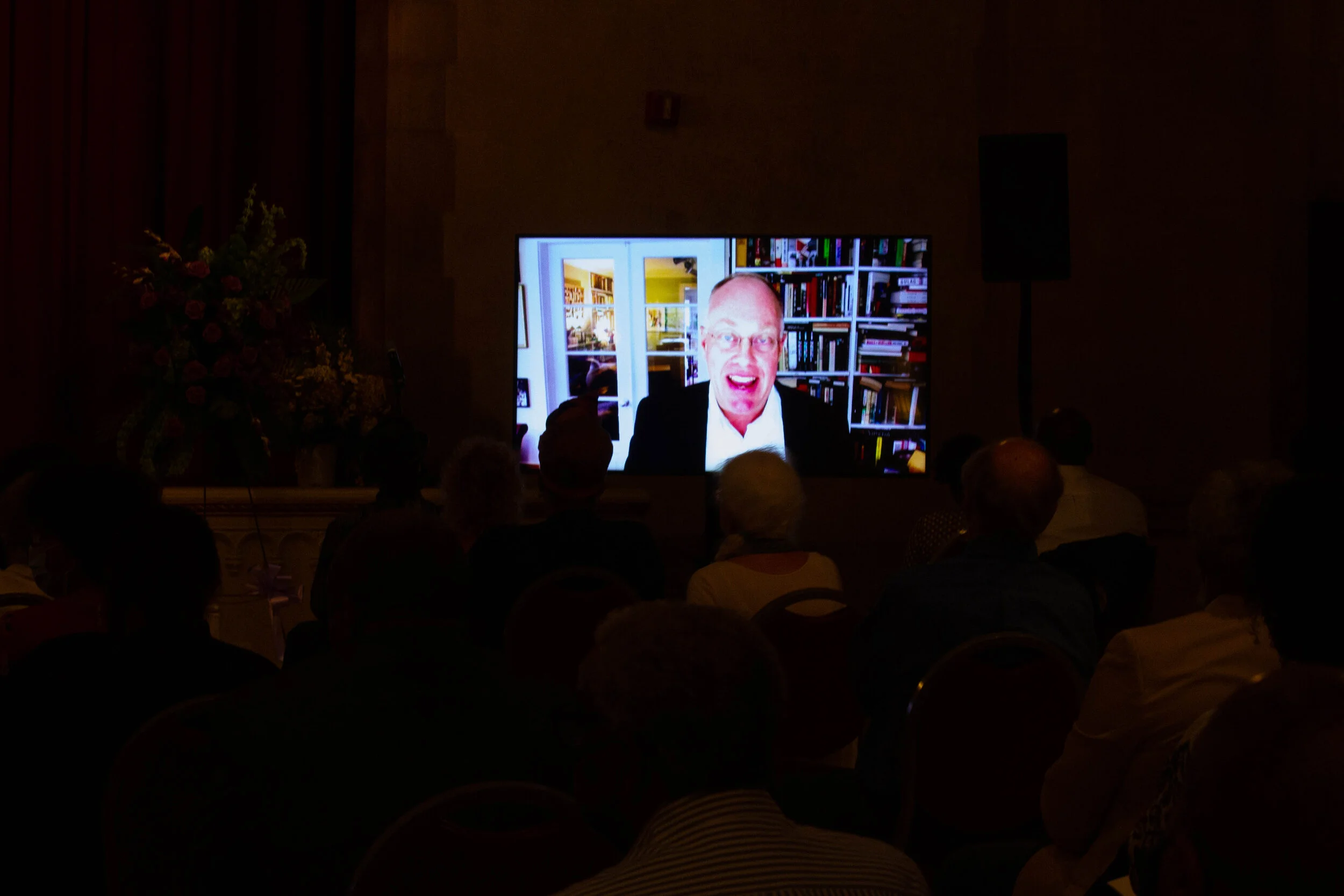 A man speaking on a video call is displayed on a large screen at an event. The background shows bookshelves and a bright room with furniture.