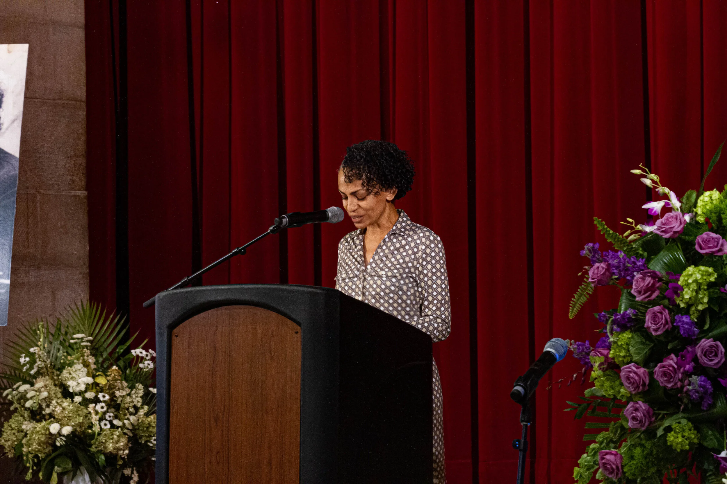 A woman with curly hair standing at a podium, speaking or reading, in front of a red curtain, with colorful flower arrangements on either side.