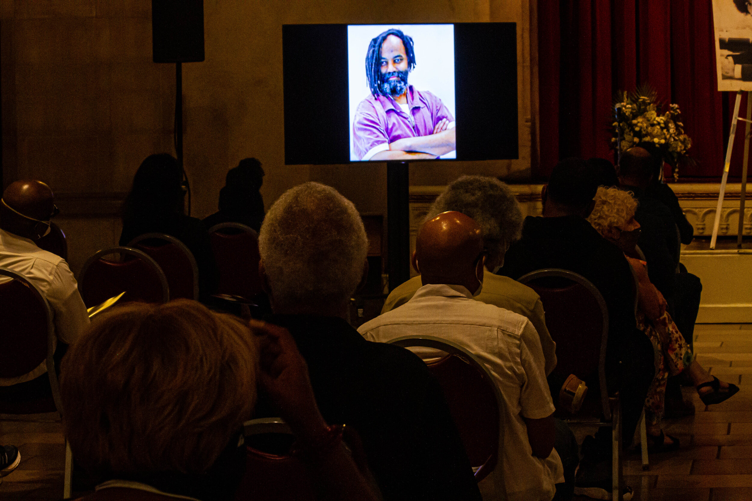 Audience seated in a dark conference room watching a large screen displaying a portrait of a man with dreadlocks and a beard, crossing his arms and smiling.