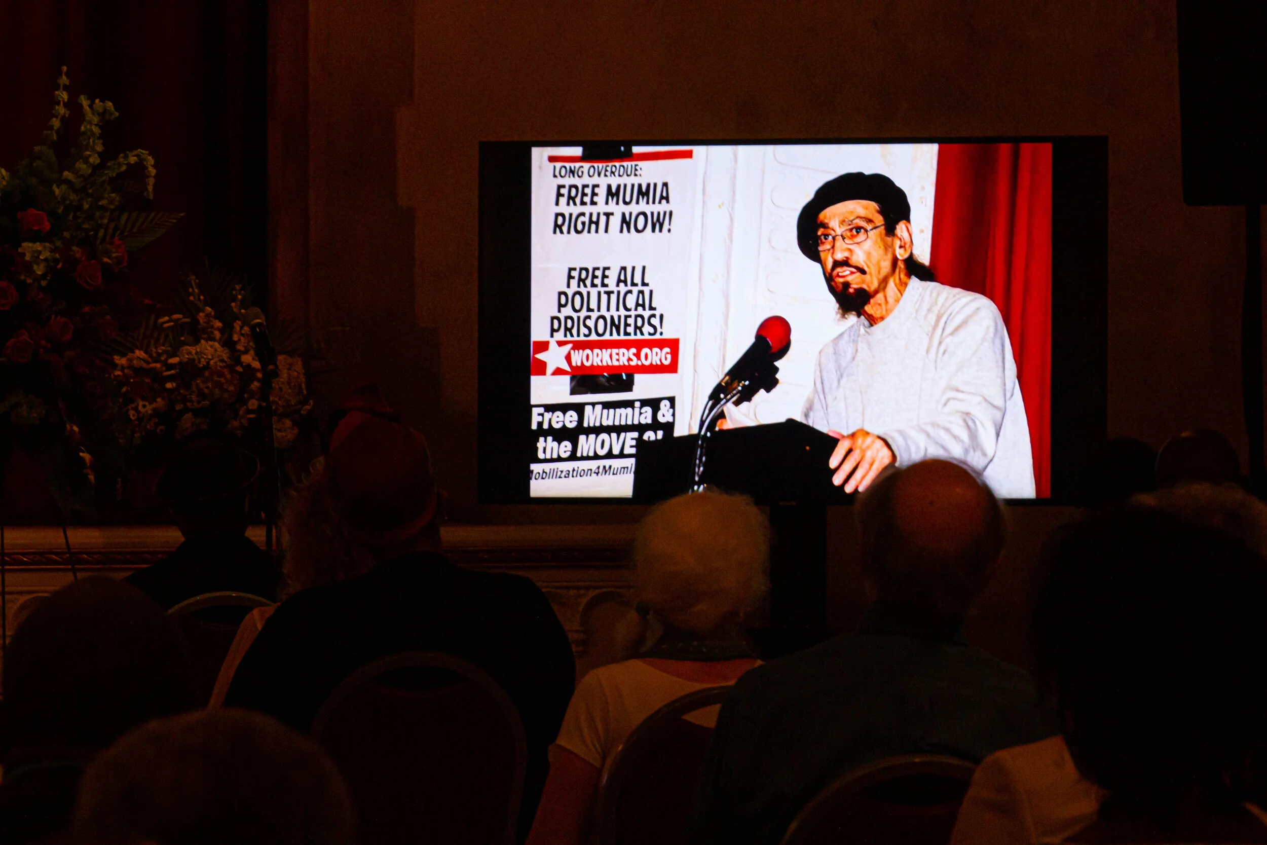 A man speaking at a podium during a conference or rally with a large screen behind him displaying a message supporting political prisoners and free Mumia. The screen includes a website URL, workers.org.