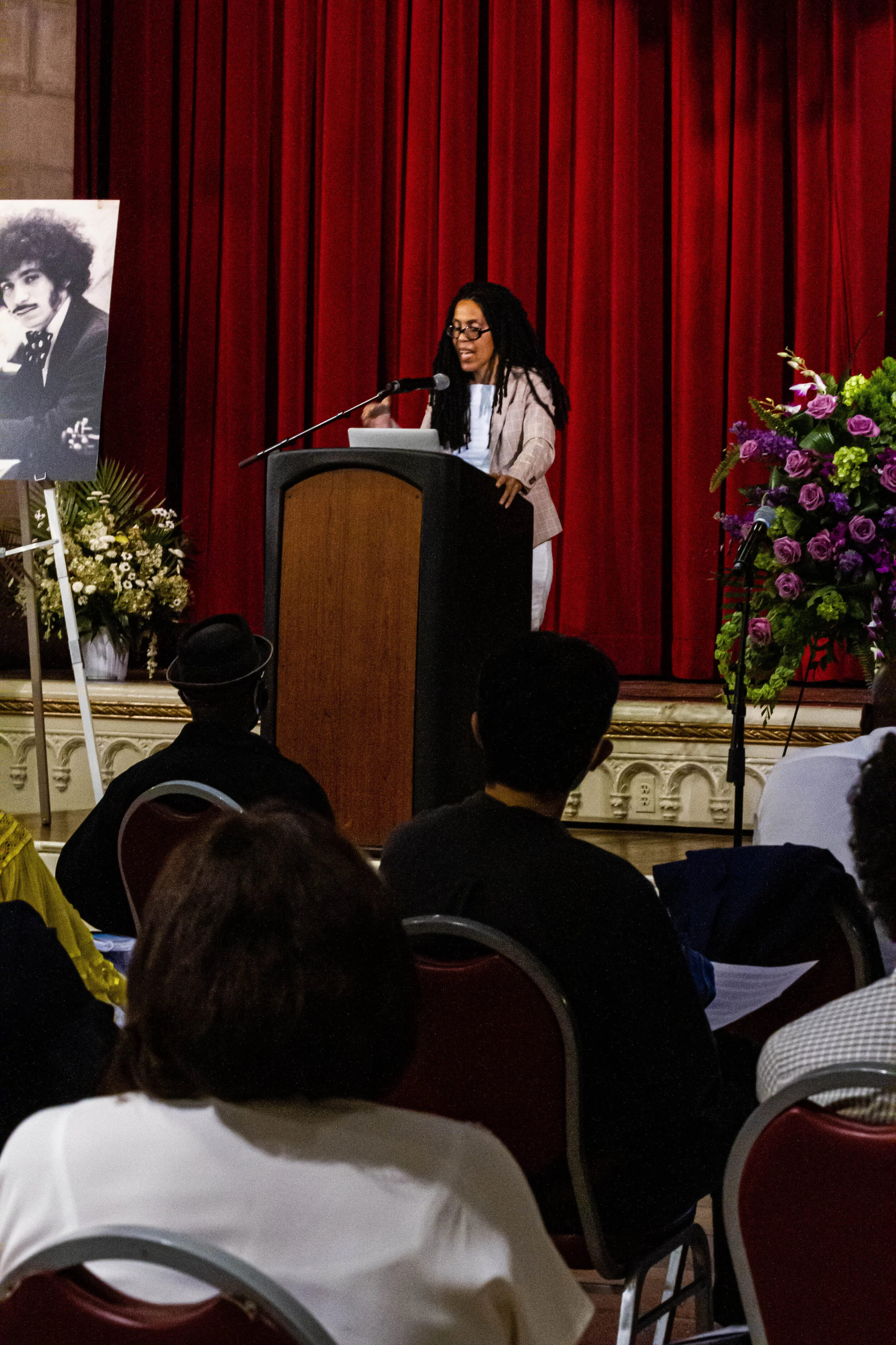 A woman with long dreadlocks and glasses, dressed in a light-colored blazer and white blouse, standing at a podium with a microphone, speaking to an audience. Behind her is a red curtain, to her right is a large bouquet of purple and green flowers, a