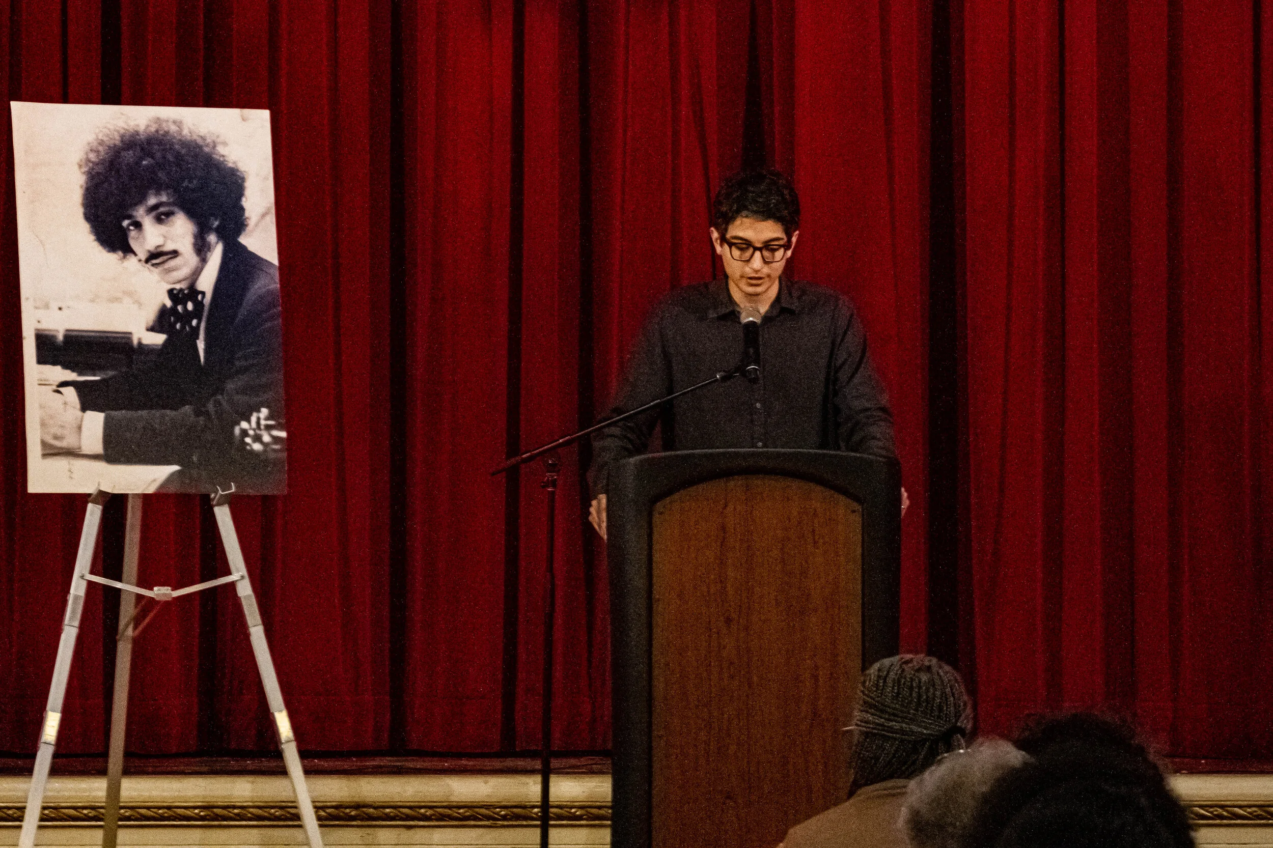 A person with glasses and dark hair speaking at a podium in front of red curtains. To the left, there is a portrait of a man with curly hair and a mustache playing the piano, displayed on an easel.