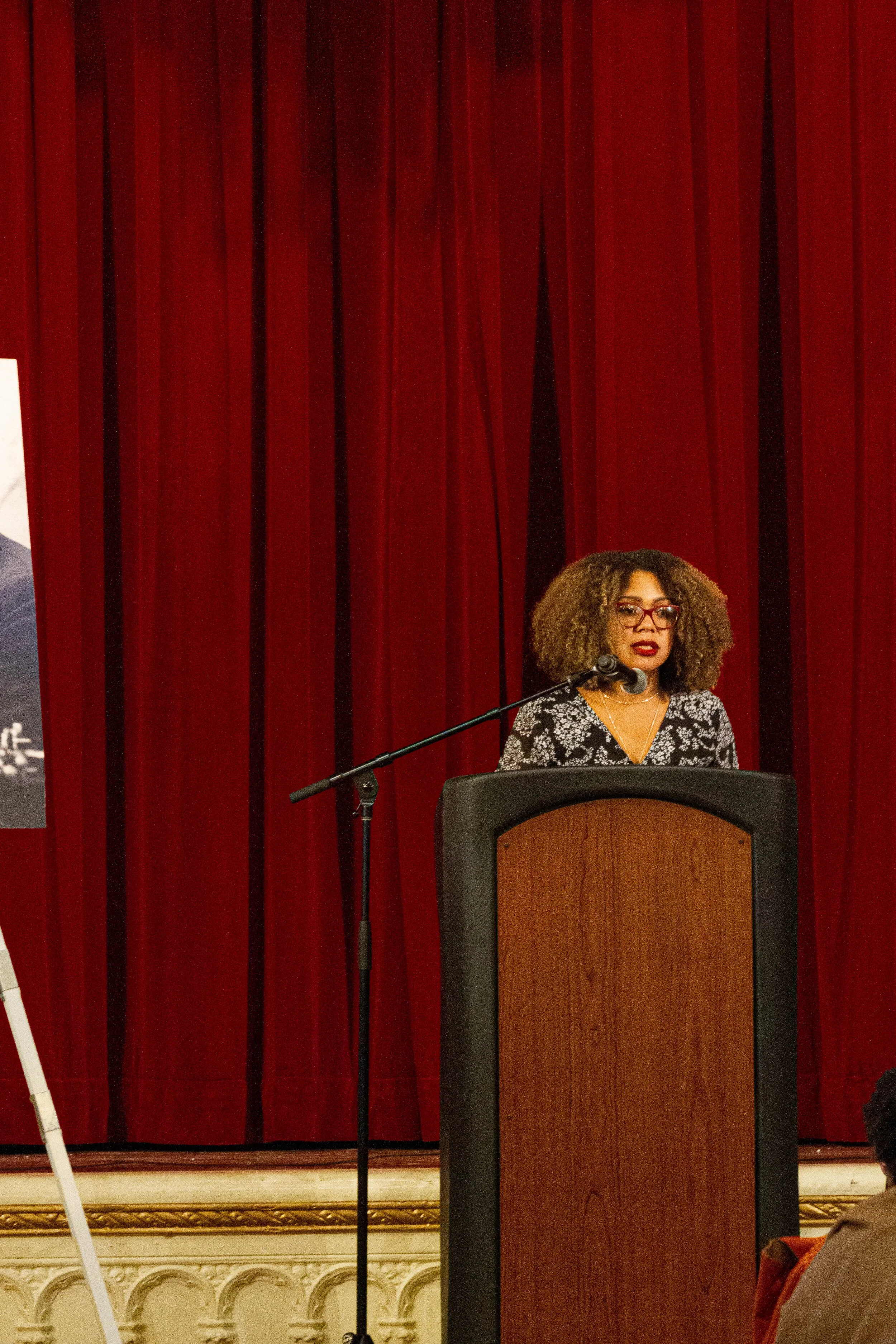 A woman with curly hair and glasses standing at a podium with a microphone in front of red curtains.