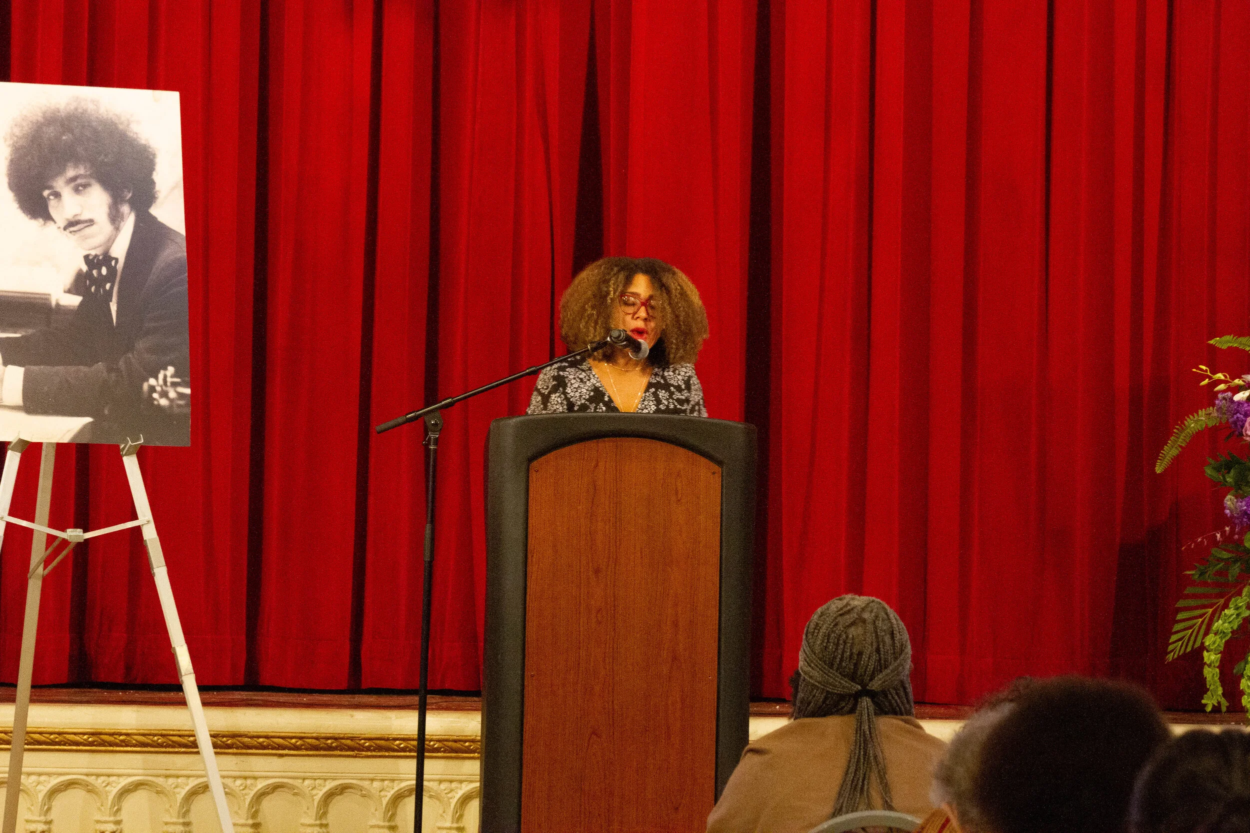 A woman with curly hair and glasses is speaking at a podium in front of red curtains. There is a large black-and-white photo of a man with an afro hairstyle and mustache displayed on an easel to her left. A small bouquet of flowers is visible on the 