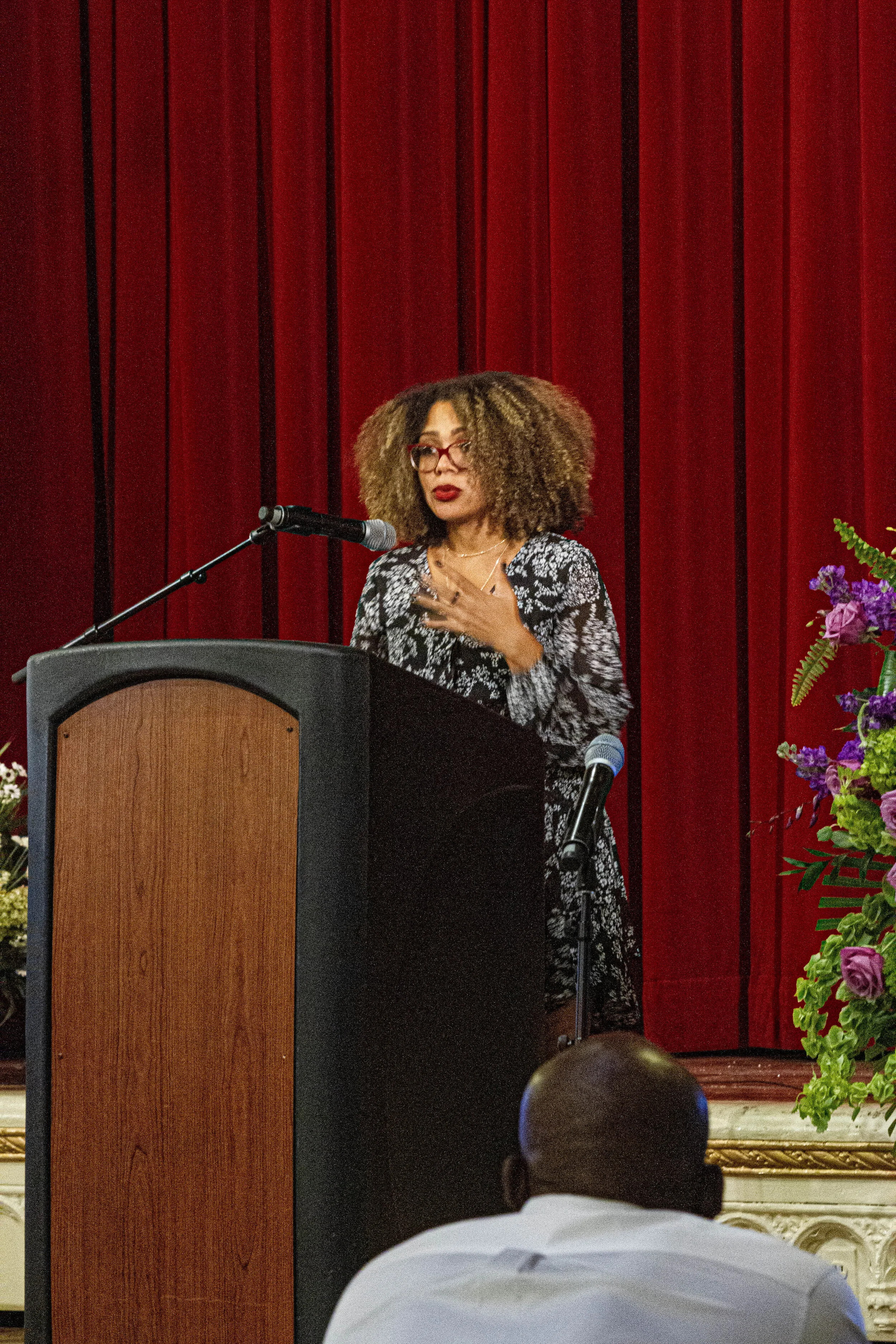 A woman with curly hair, wearing glasses and a patterned black and white dress, is standing at a podium with a microphone, speaking. She is gesturing with her right hand, and there are red curtains and a floral arrangement nearby. The audience is vis