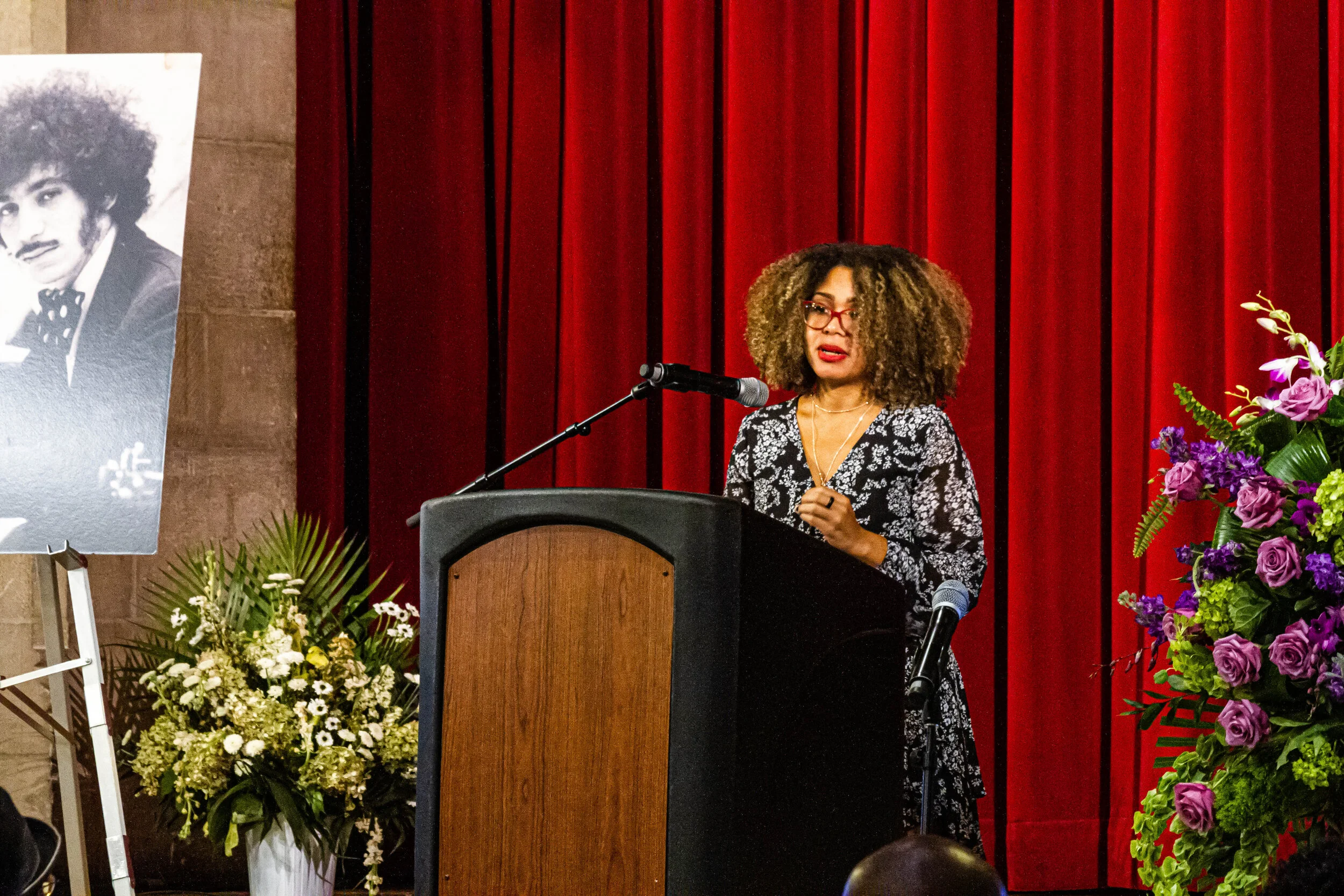Woman with curly hair and glasses speaking at a podium during a formal event. There are vibrant purple and pink flowers to her right, behind a red curtain, with a black and white portrait of a man on an easel to her left.