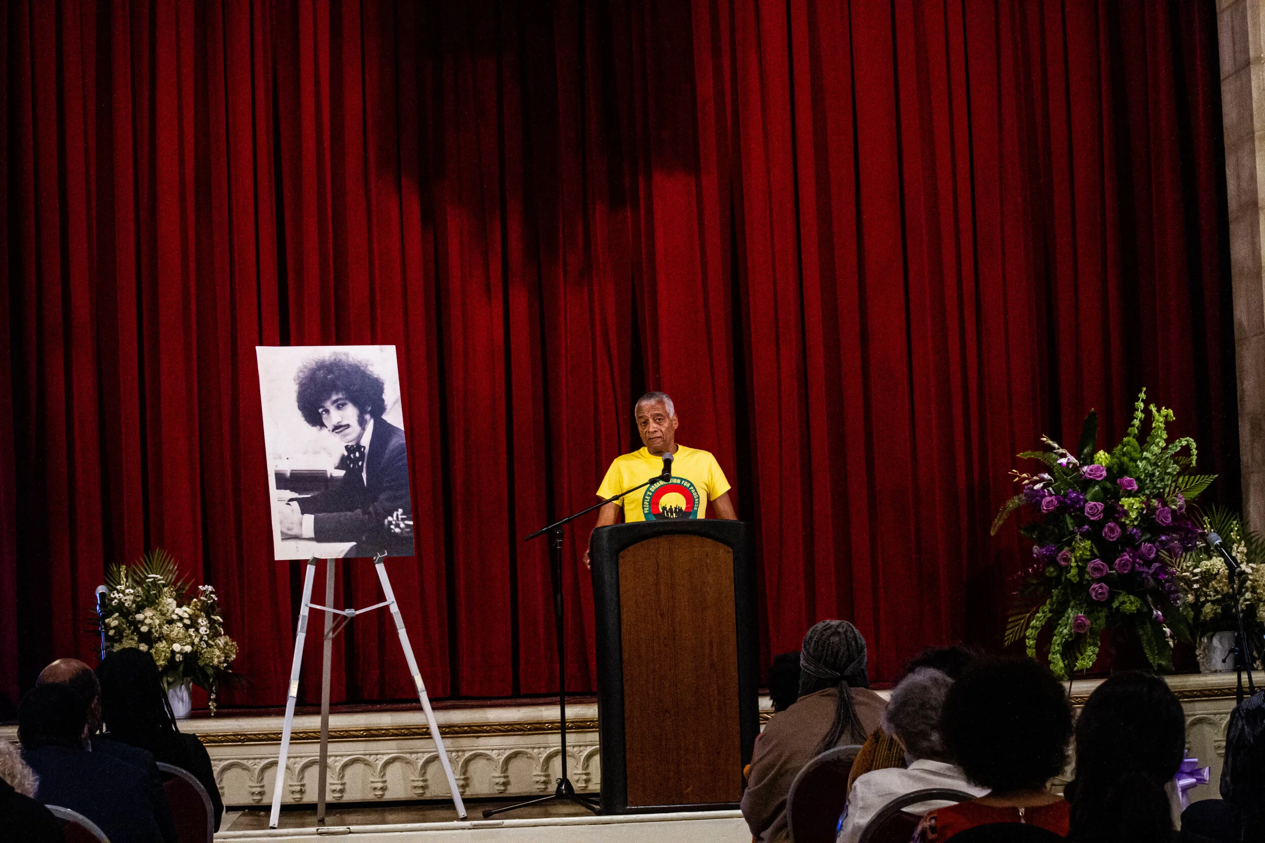 A man in a yellow shirt stands at a podium on a stage with red curtains behind him, speaking to an audience. To his left is a large portrait of a person with curly hair playing a piano, and there are floral arrangements on stage.