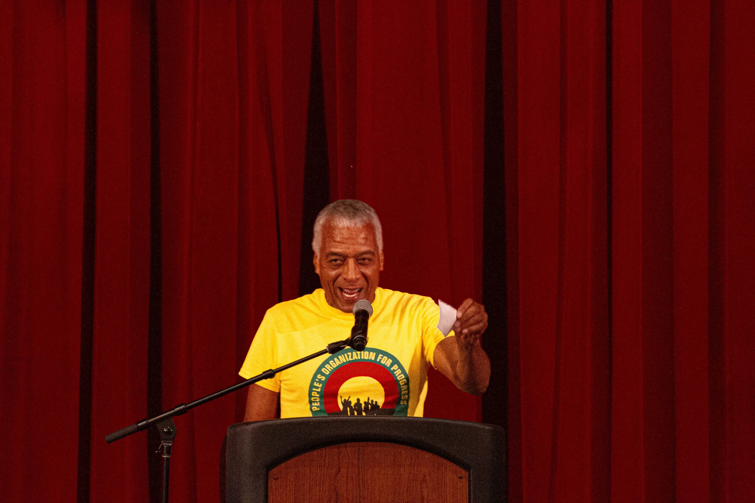 An older man with gray hair speaking at a podium with a microphone, wearing a yellow T-shirt with a logo, in front of a red curtain.