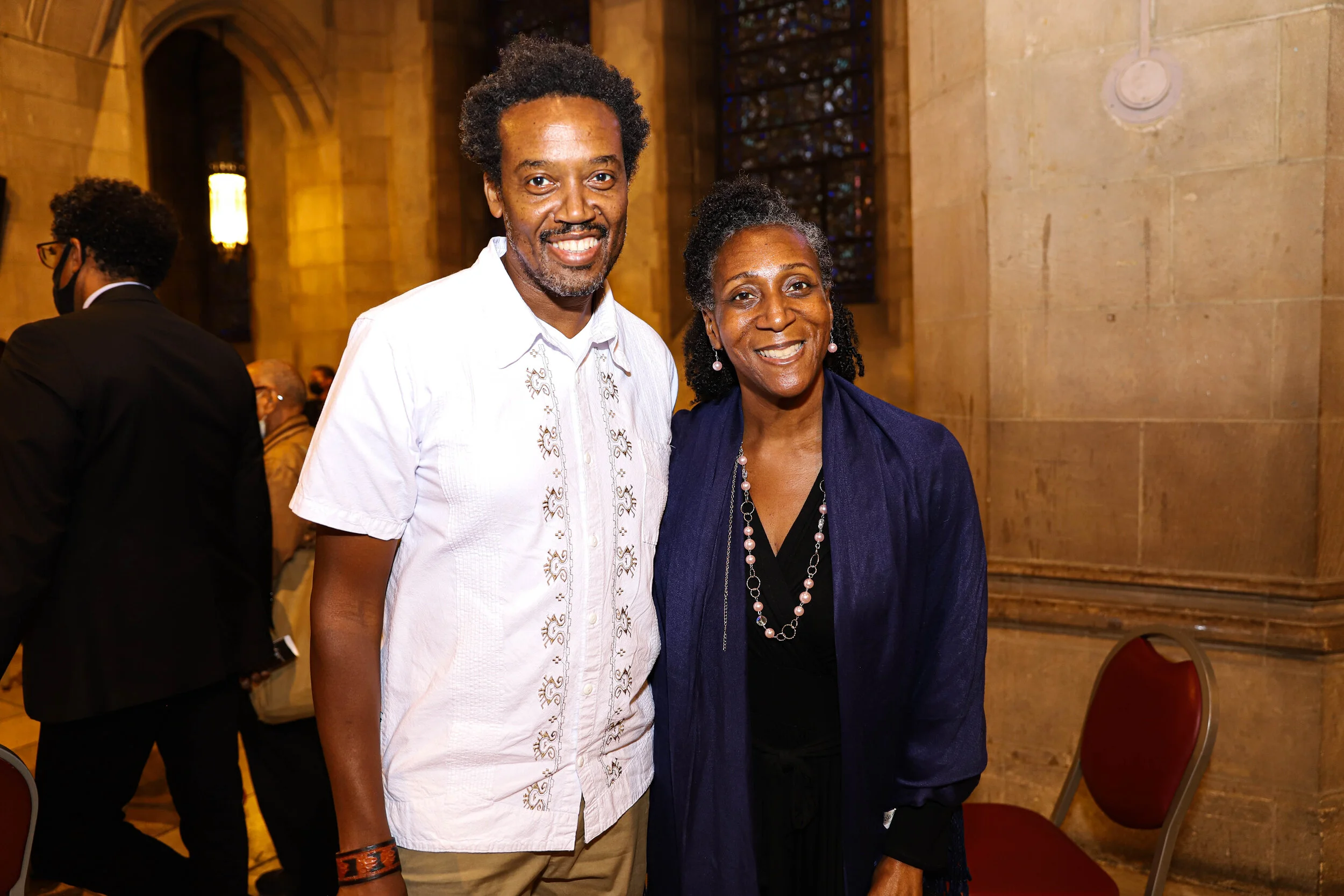 A man and a woman standing inside a church, smiling at the camera. The man is wearing a white embroidered shirt, and the woman is wearing a dark jacket with a pearl necklace. Other people are visible in the background, with stained glass windows on t