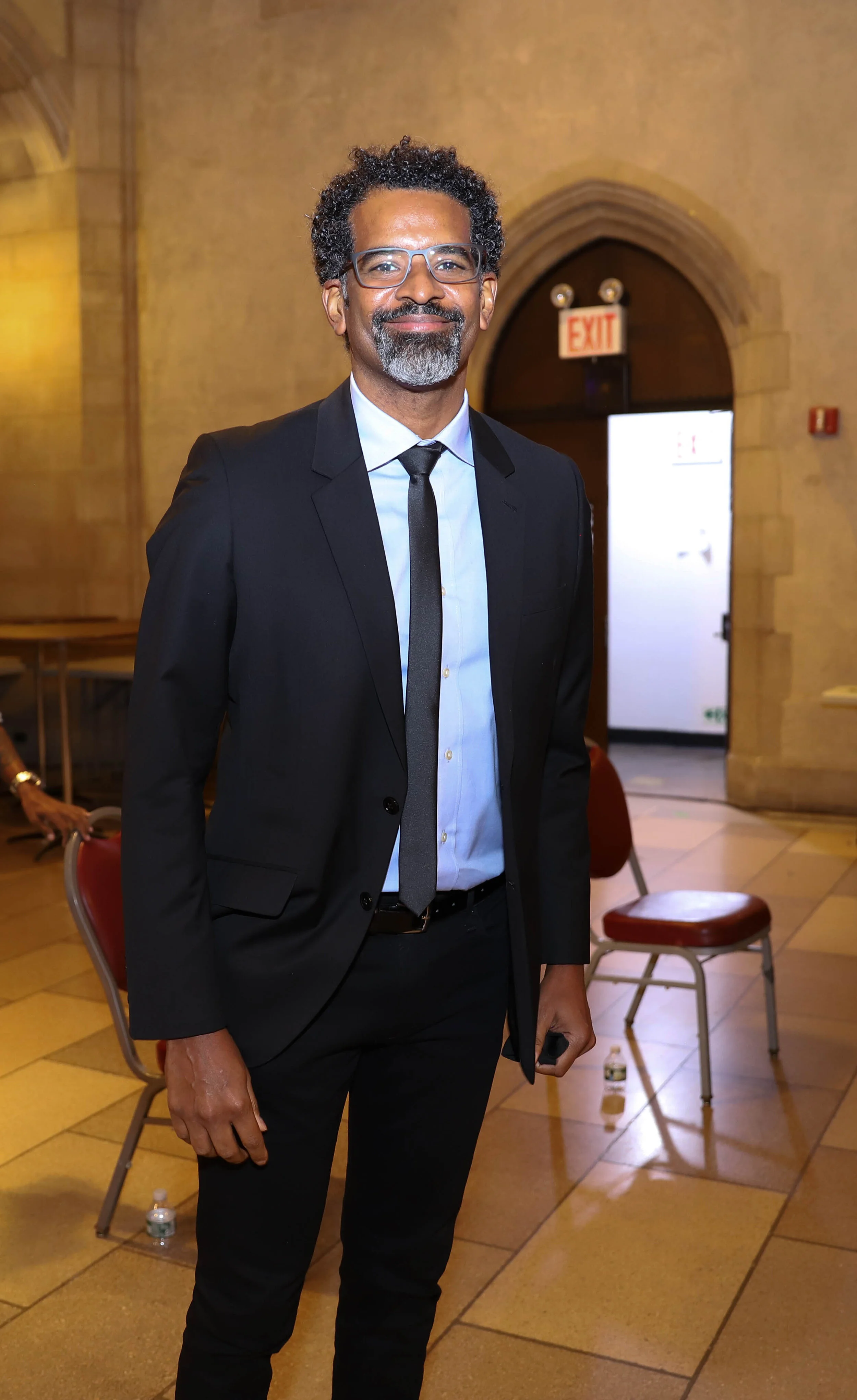 A man in a black suit, white shirt, and black tie standing indoors with chairs and an "EXIT" sign behind him.