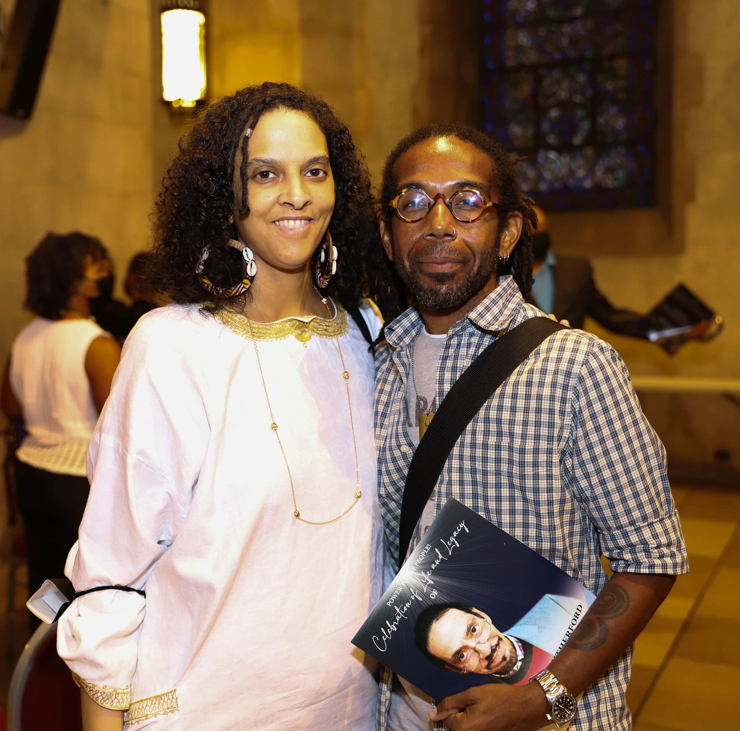 A woman with long curly hair and large shell earrings standing next to a man with dreadlocks, glasses, and tattoos, holding a magazine with a person's face on the cover at an indoor event.