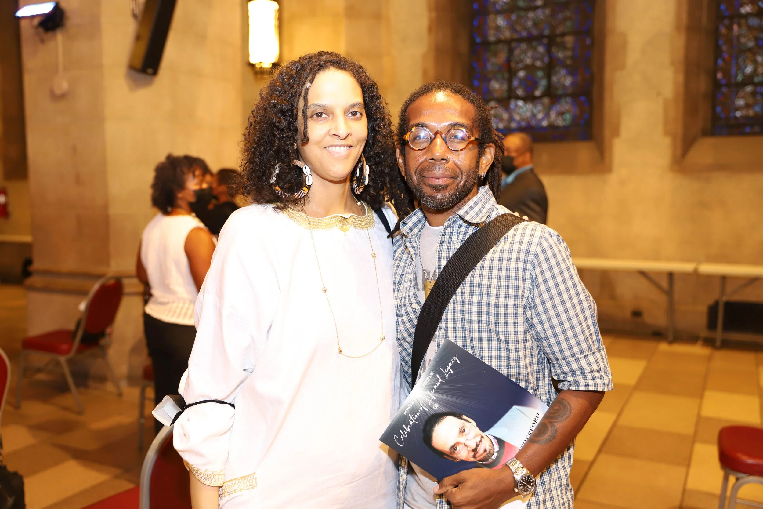 Two individuals, a woman with curly hair and large earrings and a man with glasses and dreadlocks, standing inside a church or hall with stained glass windows, holding a magazine featuring a man on the cover.