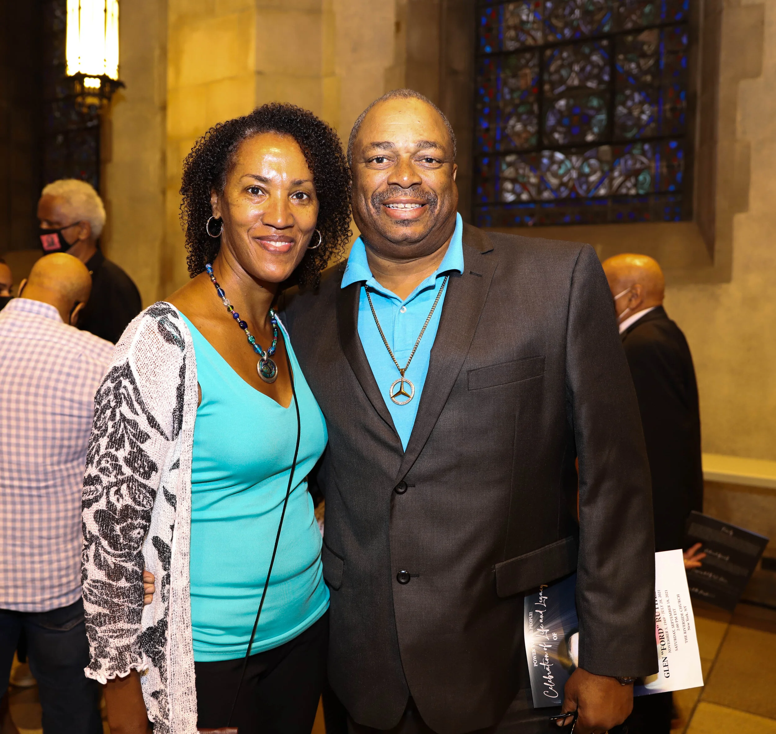 A smiling woman with curly hair and hoop earrings stands next to a smiling man with a goatee, both dressed in formal attire, in a church or cathedral setting with stained glass windows in the background.