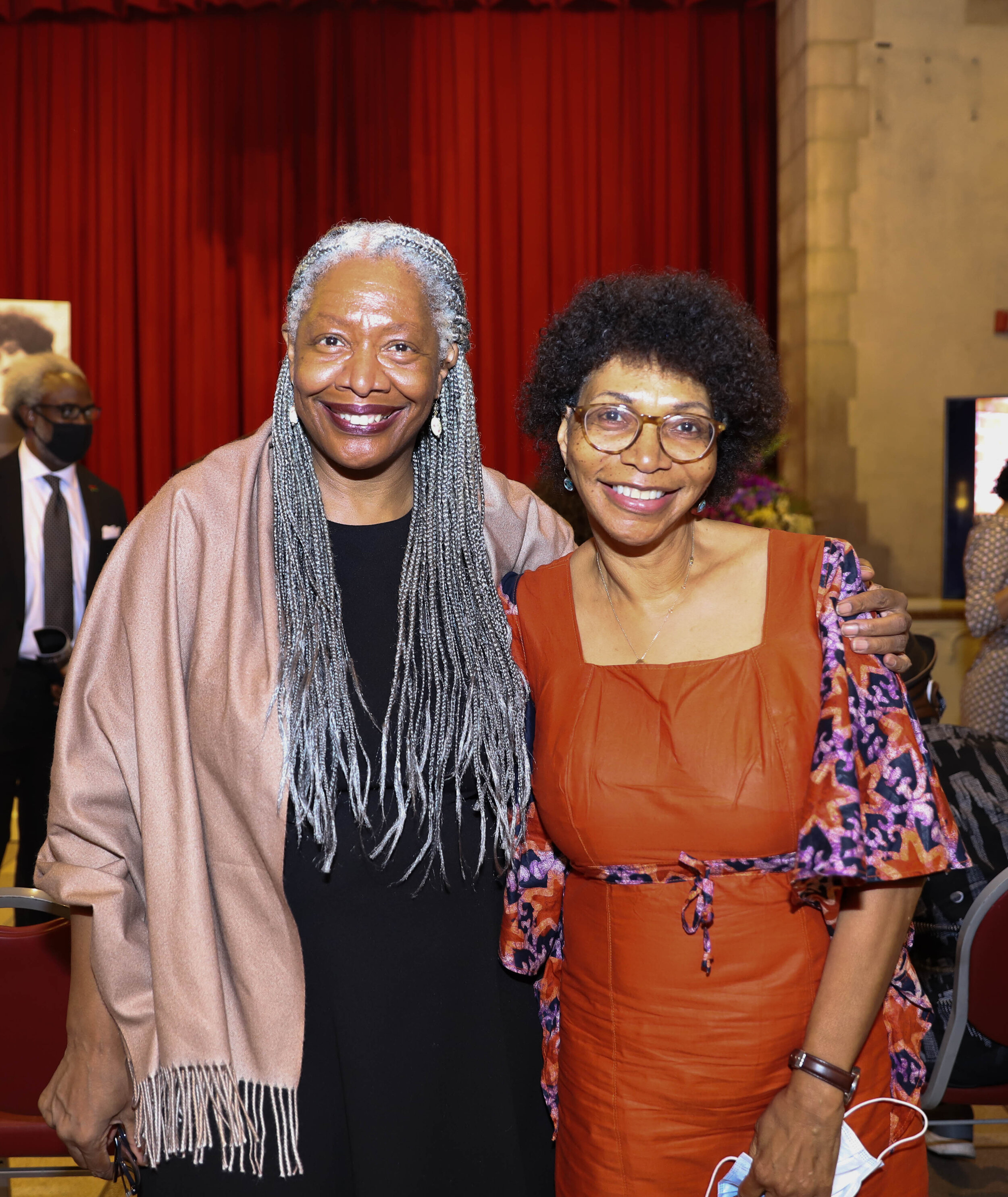 Two women smiling and posing together at an indoor event, with red curtains in the background.
