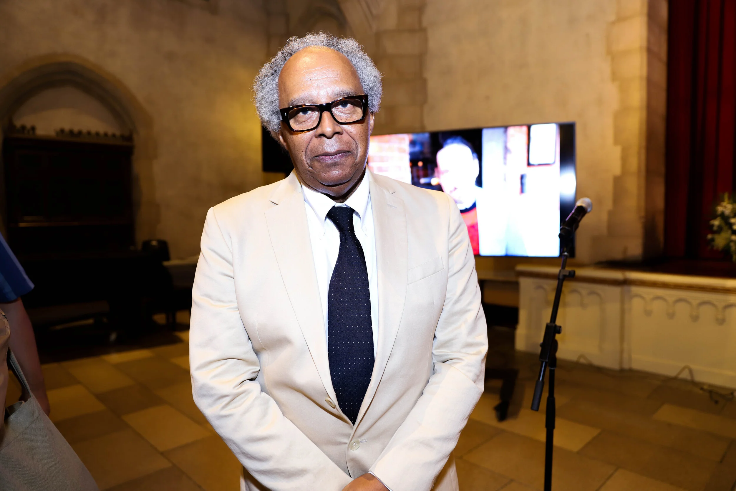 An elderly man with gray curly hair, black glasses, wearing a cream-colored suit with a white shirt and black tie, standing in a room with a large screen and red curtains in the background.