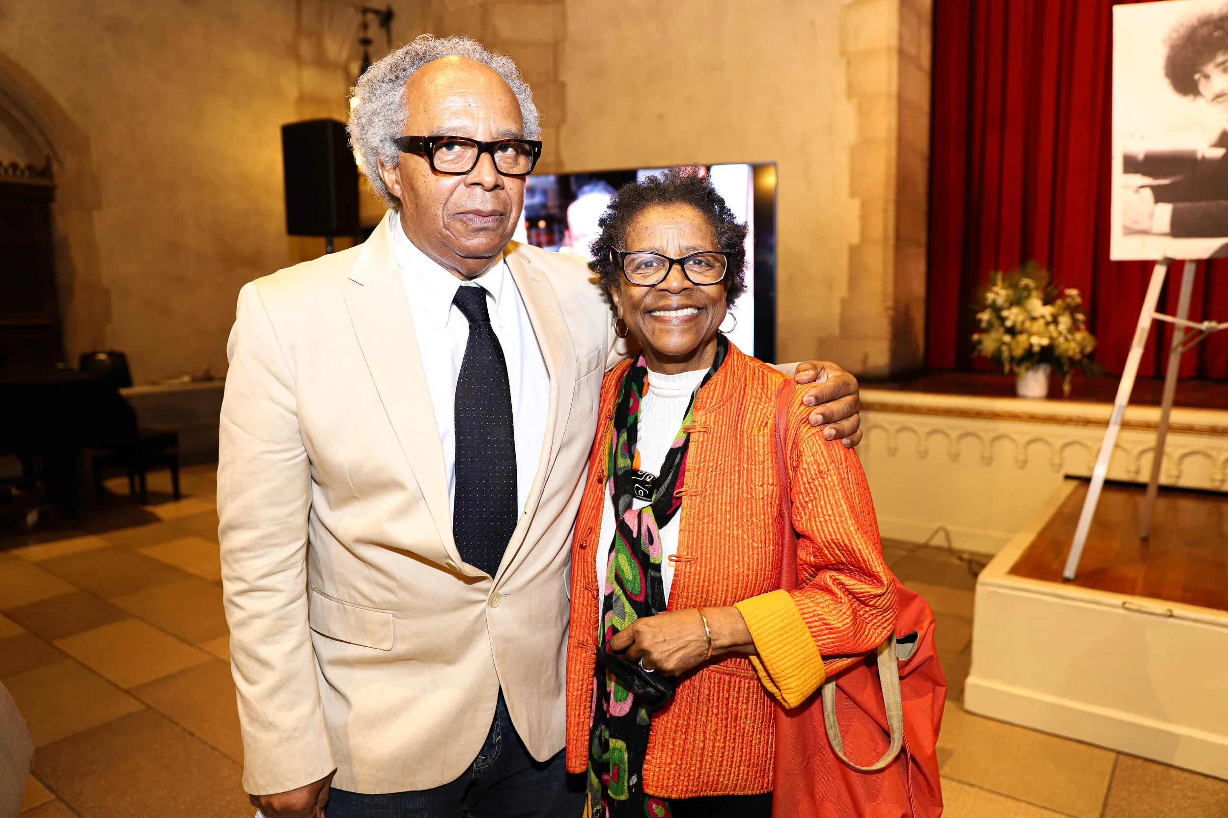 An elderly man in a beige suit, white shirt, black tie, and black glasses standing next to an elderly woman in an orange jacket, colorful scarf, and glasses. They are smiling and posing together in a formal indoor setting with a stage, flower arrange