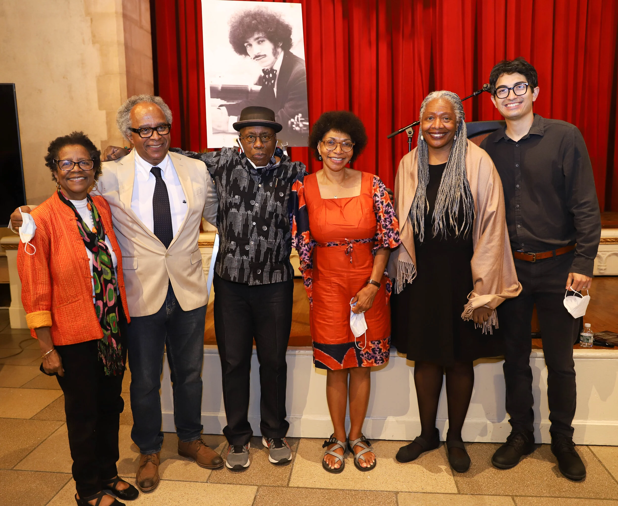 A group of seven diverse individuals standing together in front of a red curtain and a large framed black-and-white portrait of a man with curly hair and a mustache at an indoor event.