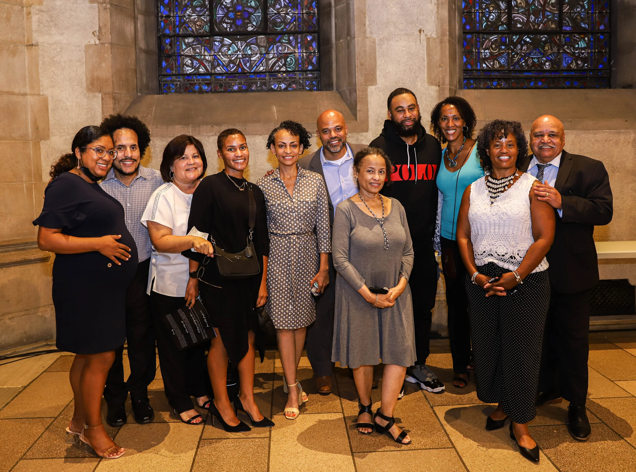 A diverse group of people, including men and women, standing together inside a building with stained glass windows in the background, smiling for a group photo.