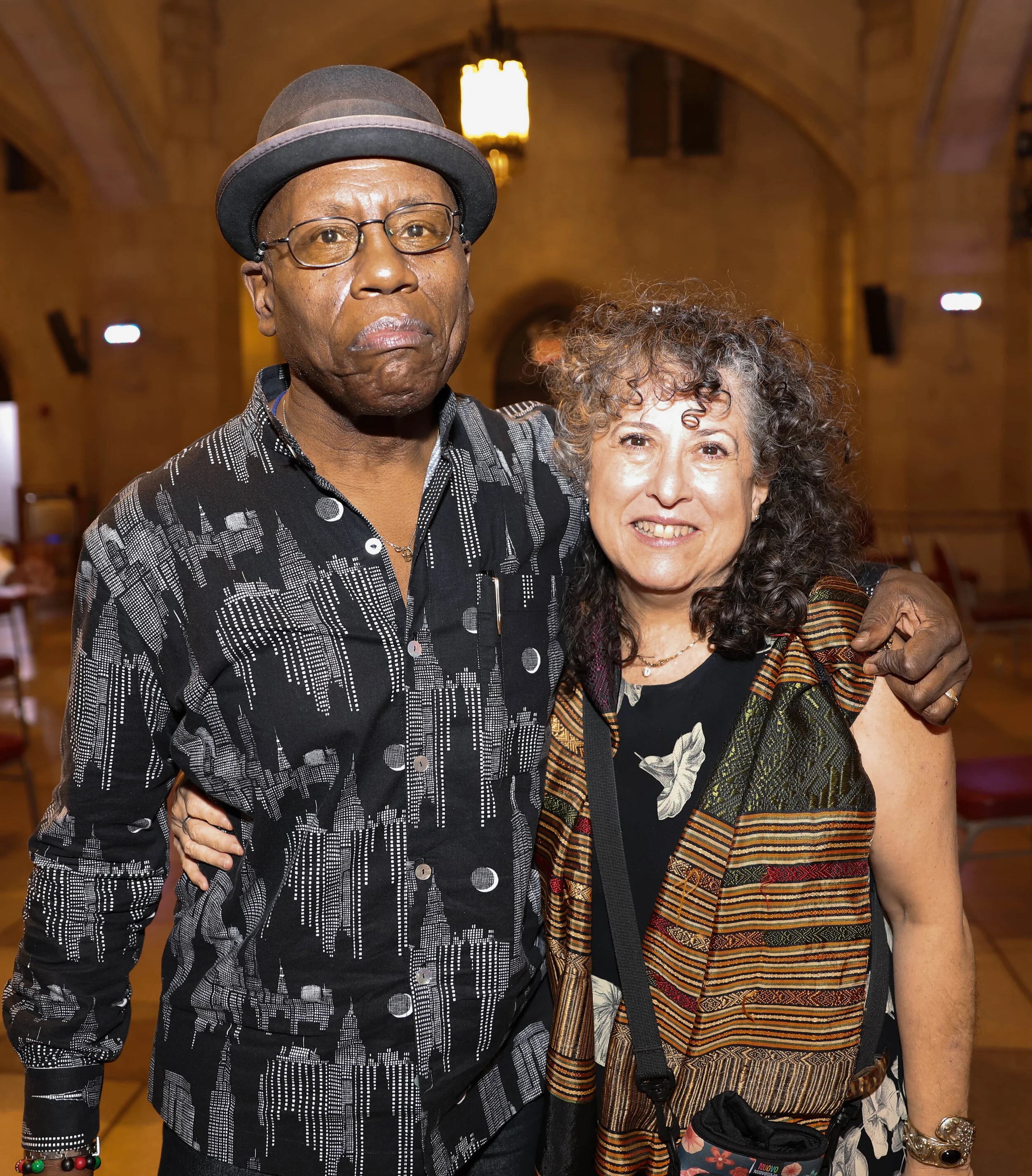 A man wearing glasses, a black hat, and a printed black and white shirt stands next to a woman with curly brown hair, wearing a colorful patterned dress, inside a warmly lit room with wooden arches.